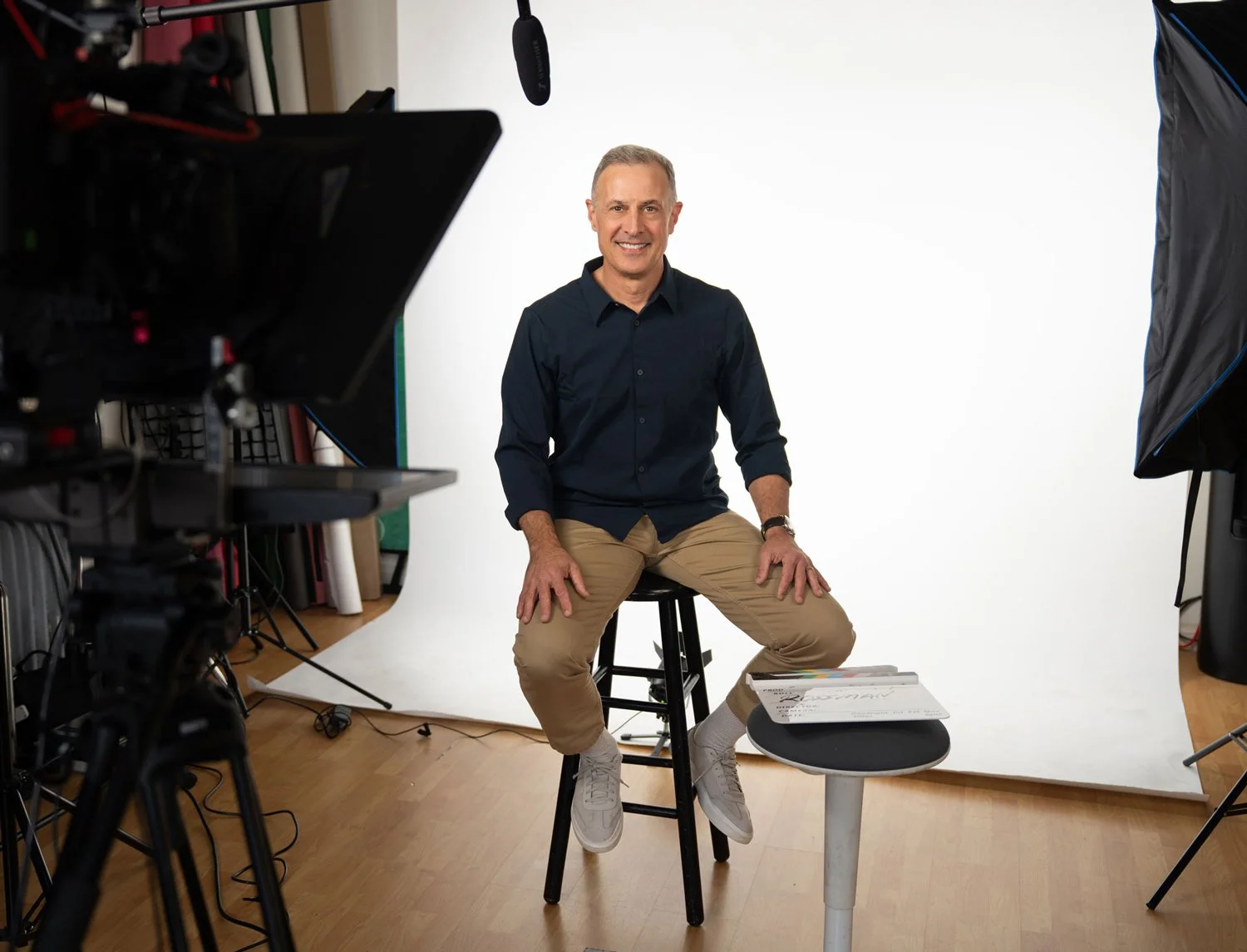 Man sitting on a stool in a photography studio, smiling at the camera, with video equipment and a white backdrop around him.