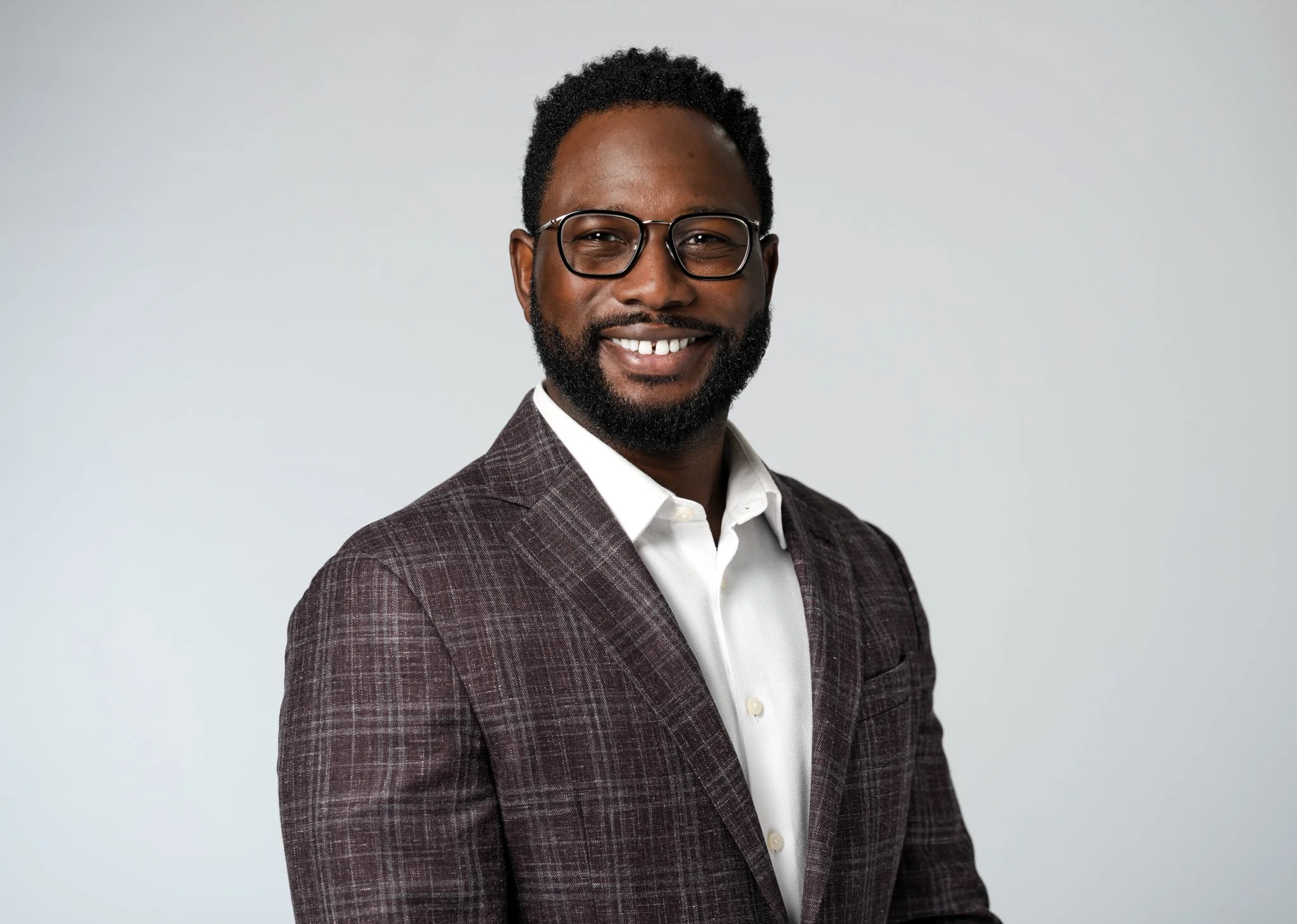 A man with glasses, a beard, and dark curly hair smiling, wearing a checkered suit jacket and white shirt against a plain light background.