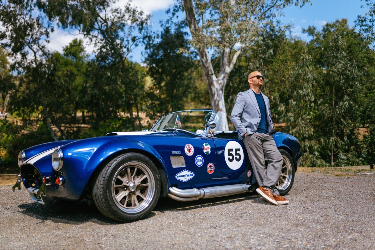 A man in a gray blazer, blue shirt, and gray pants, wearing sunglasses, standing outdoors next to a vintage blue racing car with the number 55 and various sponsor stickers, on a gravel surface with trees in the background.
