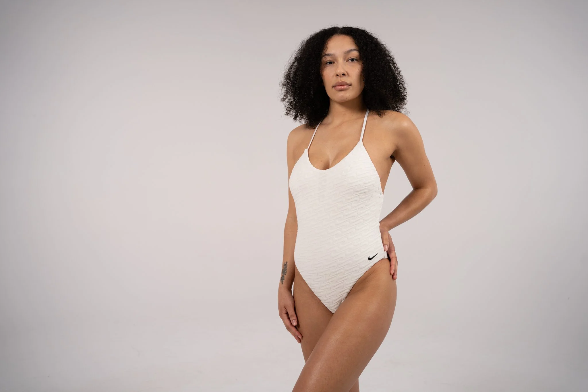A woman standing against a plain gray background, wearing a white textured swimsuit with thin straps and a small Nike logo, with her hand resting on her hip.