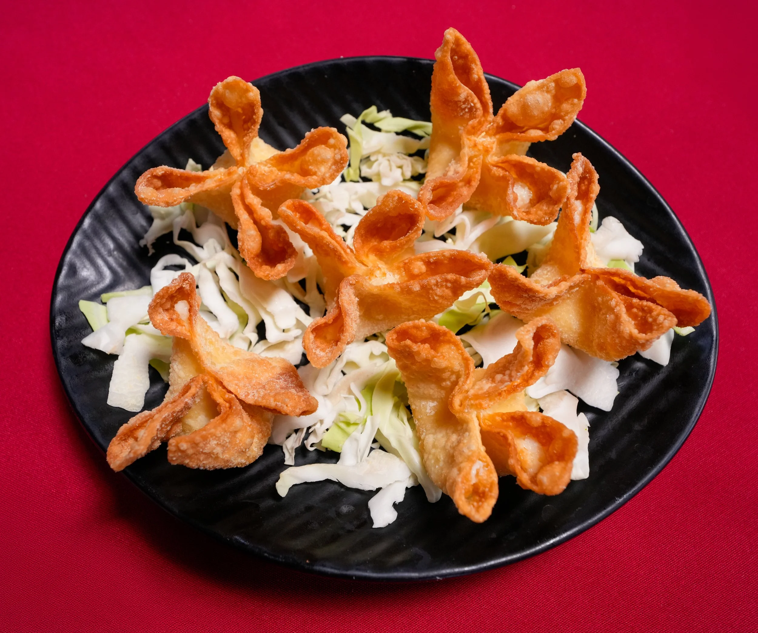 Fried wontons served on a bed of shredded lettuce in a black bowl, with a red background.