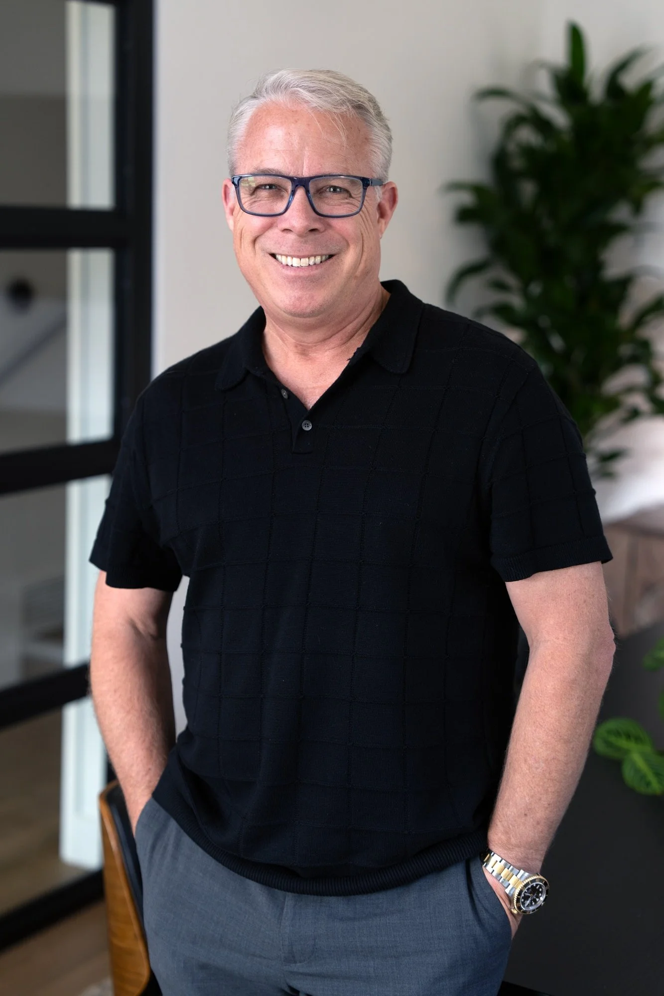 A smiling older man with gray hair and glasses, wearing a black polo shirt and gray pants, standing in an indoor setting with a plant and window in the background.