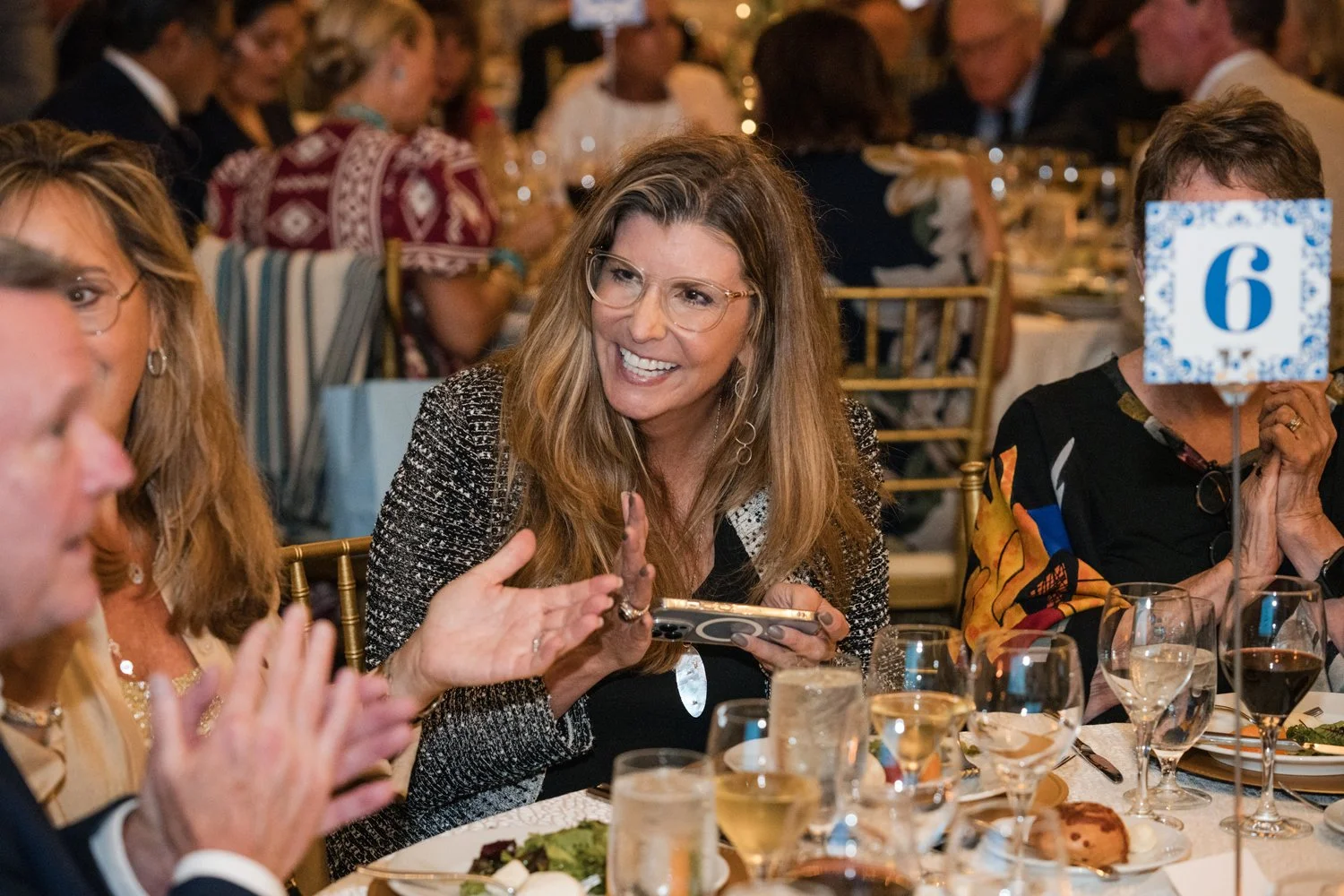 A woman with long light brown hair, glasses, and a black and white patterned blazer, smiling and engaging in conversation at a formal dinner event. She is holding a smartphone in her right hand. The table is set with wine glasses, plates of food, and