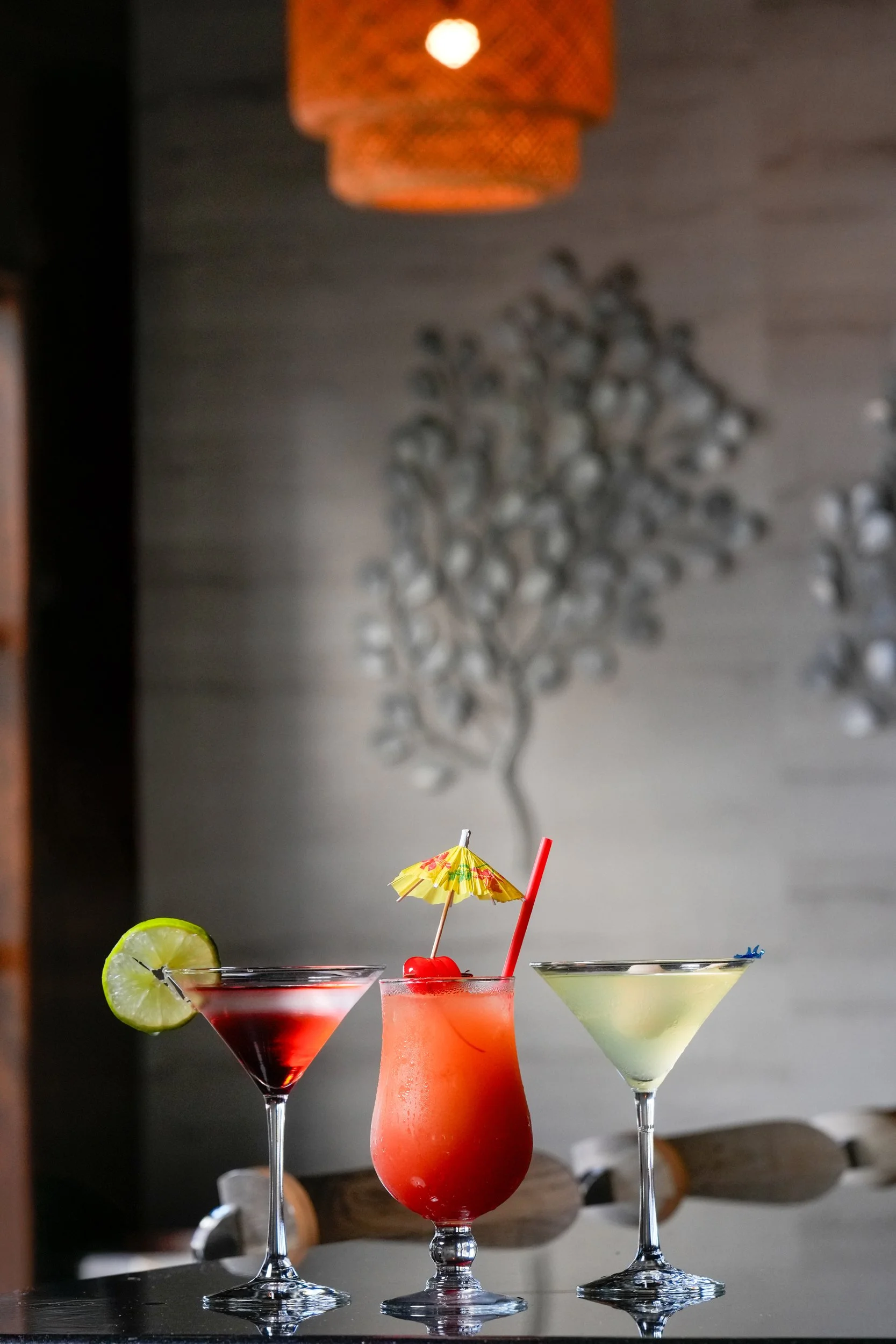 Three colorful cocktails in martini glasses with lime slice, cocktail umbrellas, and straws, set against a gray wall with a decorative shadow and a hanging orange lamp.