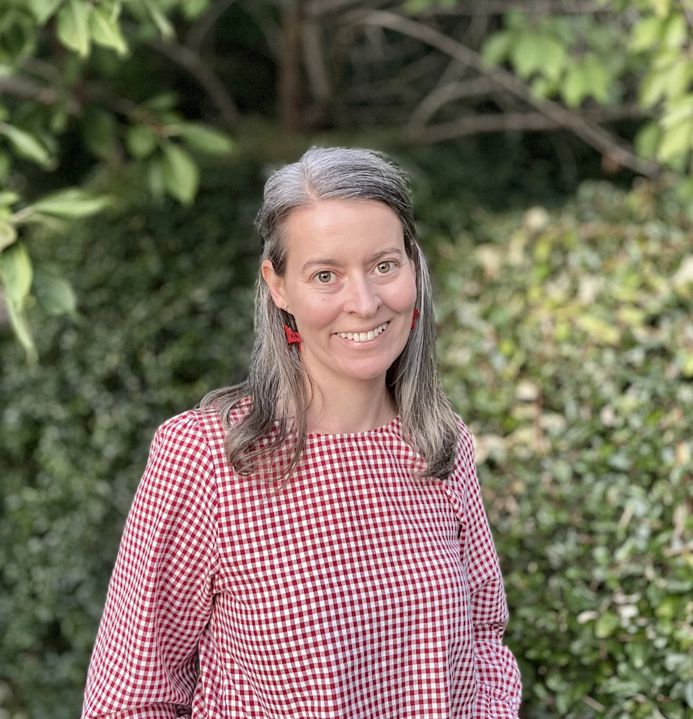 A woman with shoulder-length gray hair, wearing a red and white checkered top and red earrings, smiling outdoors with greenery in the background.