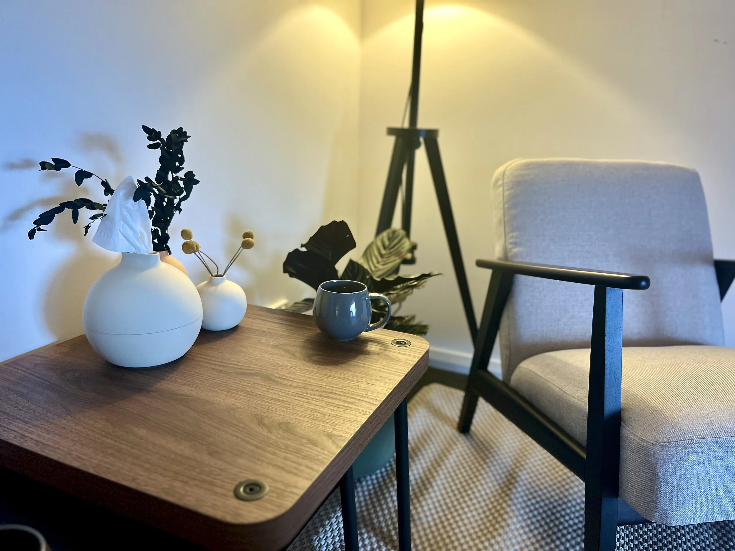 A picture of Elm Clinic interior. A wooden table with three white ceramic vases holding dried flowers and foliage, a blue mug, and a white upholstered chair next to the table. In the background, a tripod lamp.