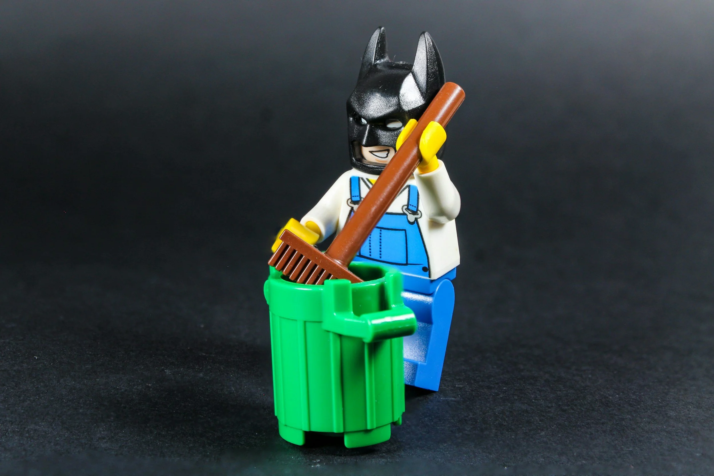 Batman is holding a broom and standing by a green trash can