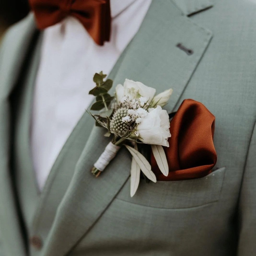 Close-up of a boutonniere with white flowers, greenery, and a white ribbon, pinned to a light gray suit jacket with a brown pocket square.