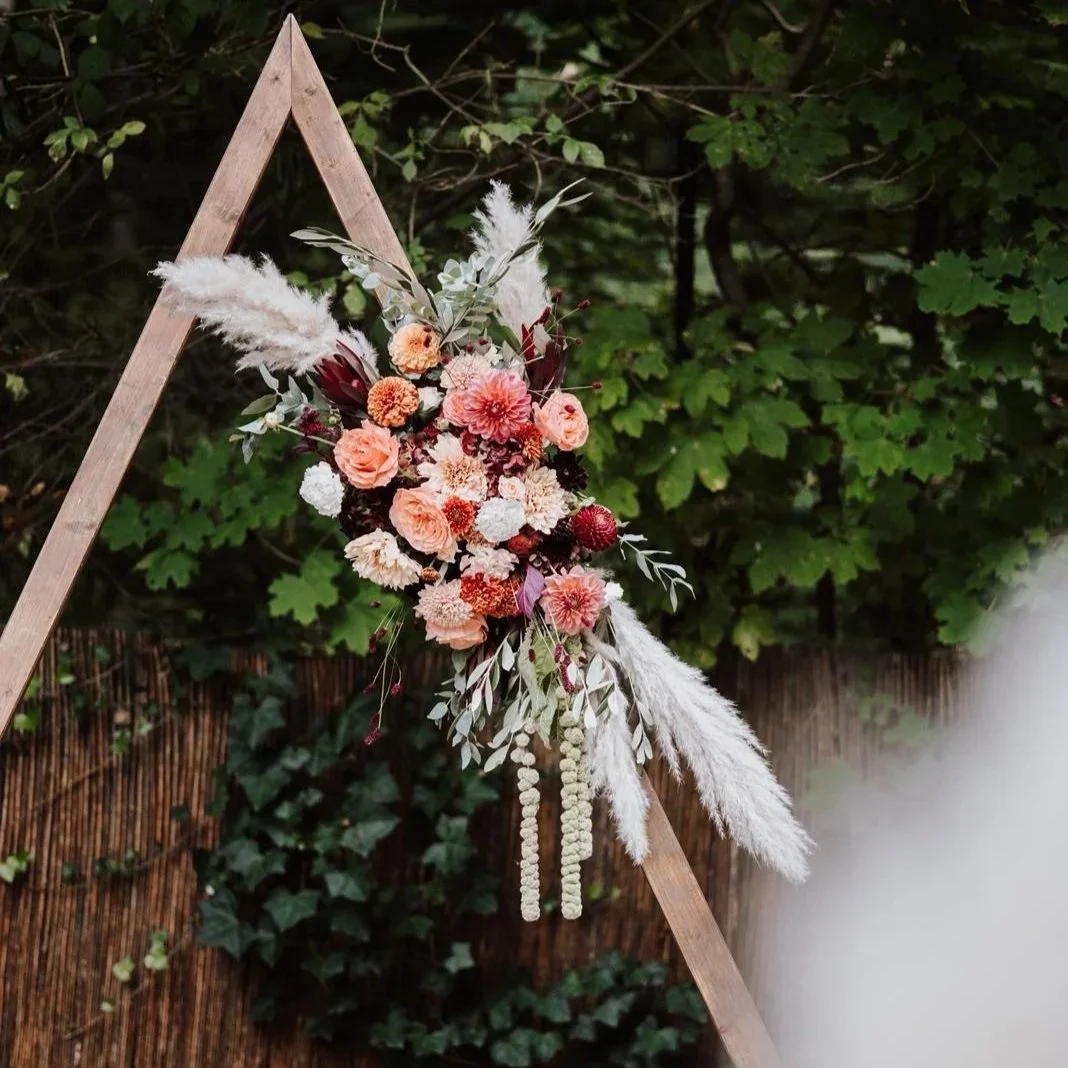 Decorative floral arrangement with pink, white, and burgundy flowers, pampas grass, and greenery on a wooden arrow-shaped frame outdoors, with lush green foliage in the background.