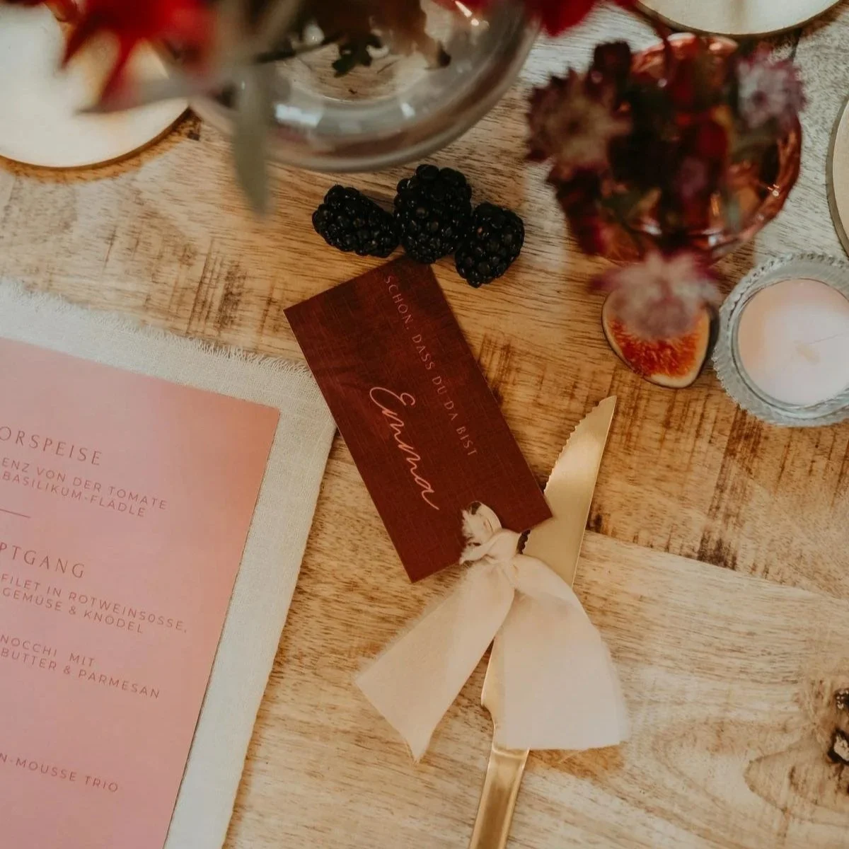 A wooden table decorated with blackberries, a pink menu, a gold-handled cake server with a cream-colored bow, a small vase with dried flowers, a candle in a glass holder, and a red card with cursive writing.