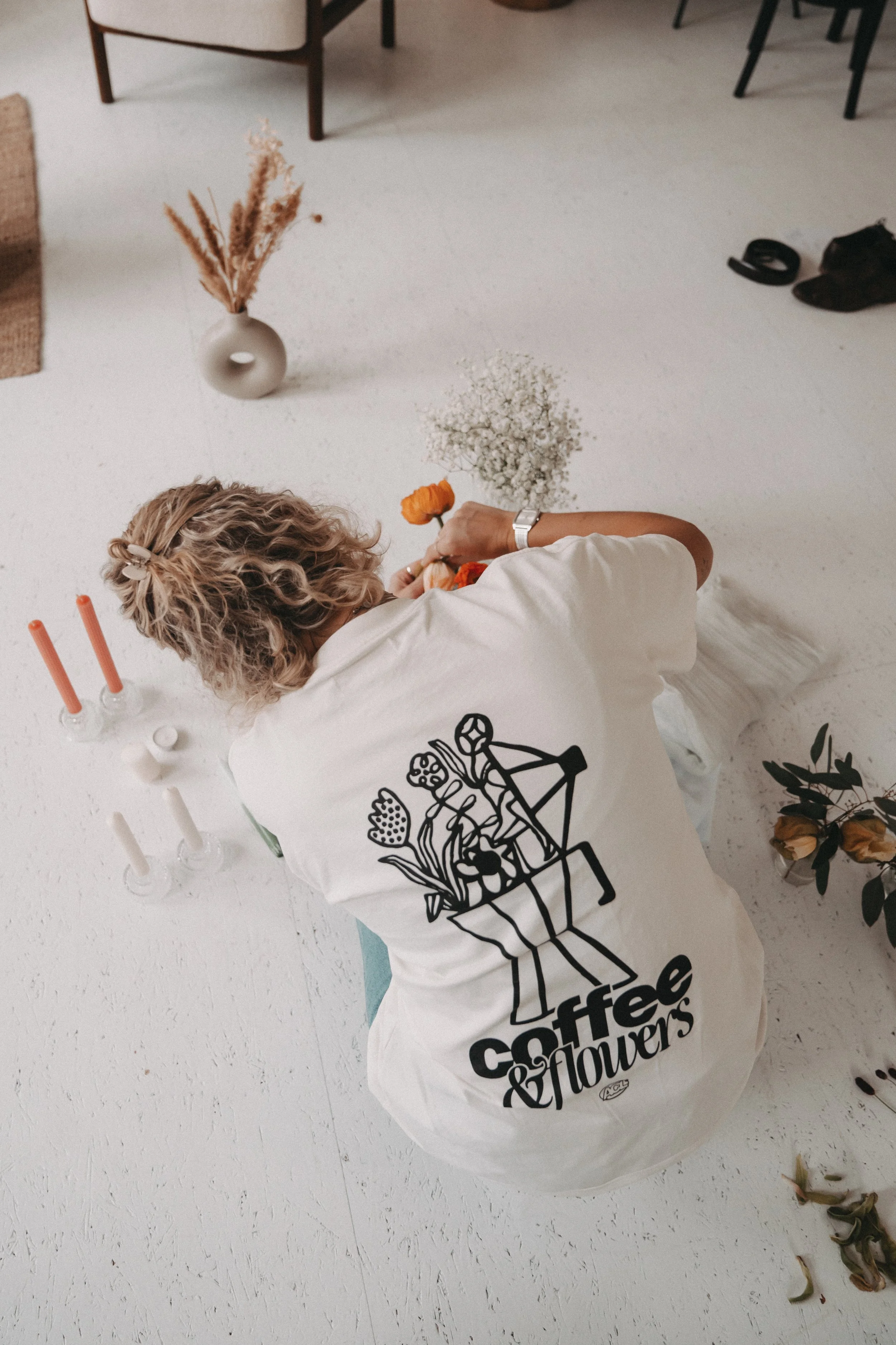 Person arranging flowers on a white floor, wearing a white T-shirt with black floral design and text 'coffee & flowers' on the back. Nearby are candles, vases with flowers, and some personal items.