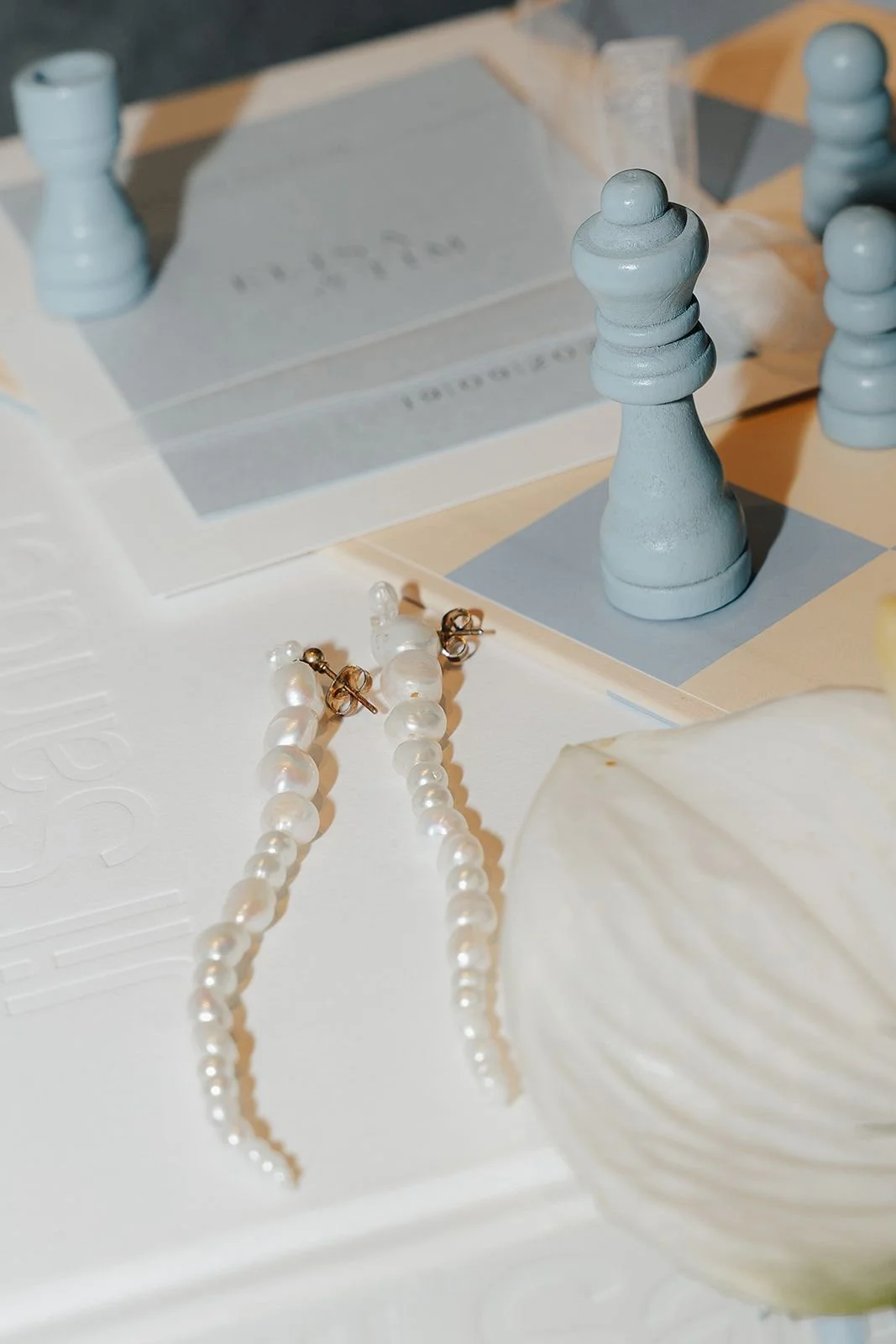 Close-up of pearl earrings, blue chess piece, and a shell on a white surface with soft lighting.