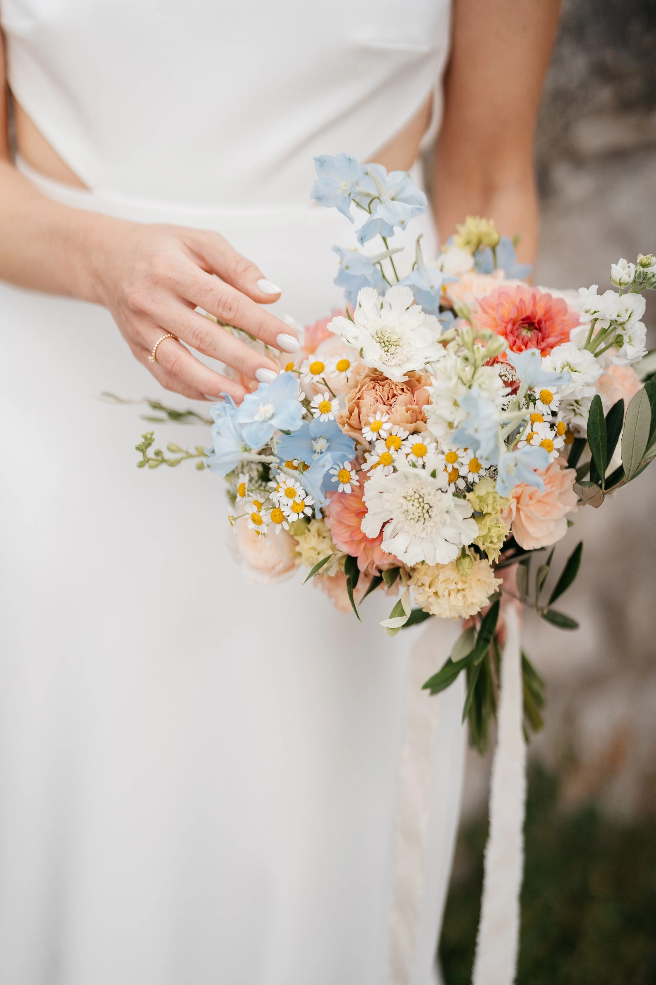 A person holding a colorful bouquet of flowers, including white, peach, and light blue blooms, with green leaves, in a white dress.