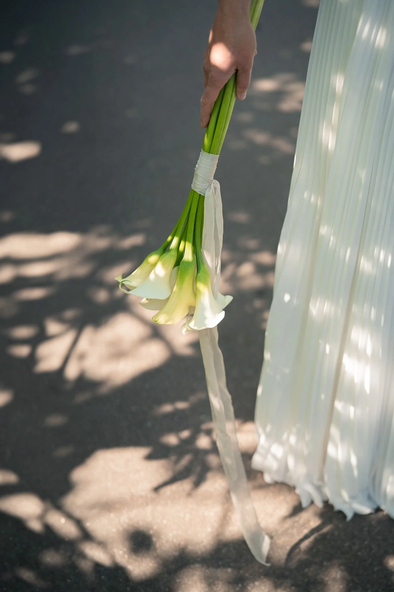 A person holding a bouquet of white calla lilies with a white ribbon, standing on a shaded pavement.