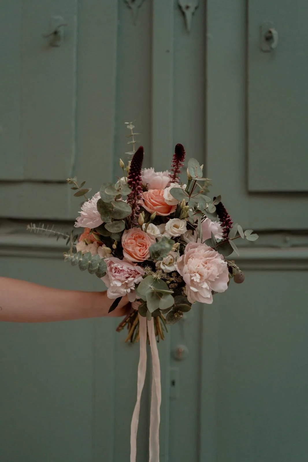 A hand holding a bouquet of pink and white flowers with greenery in front of a pale green wooden background.