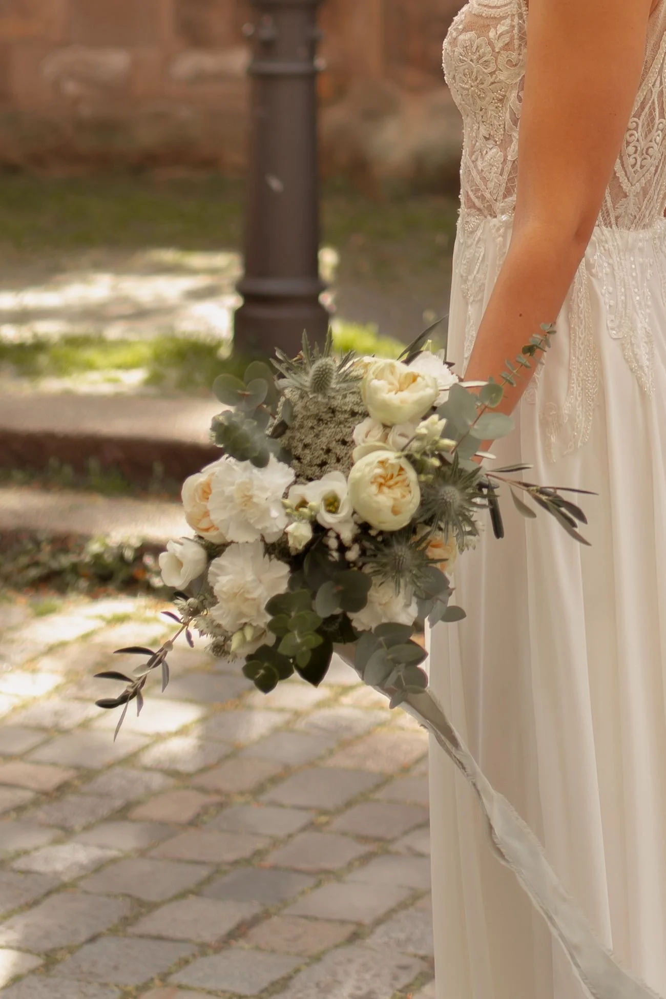 A woman in a lace wedding dress holding a bouquet of white and cream peonies, eucalyptus, and textured flowers, standing on a cobblestone path outdoors with blurred background.