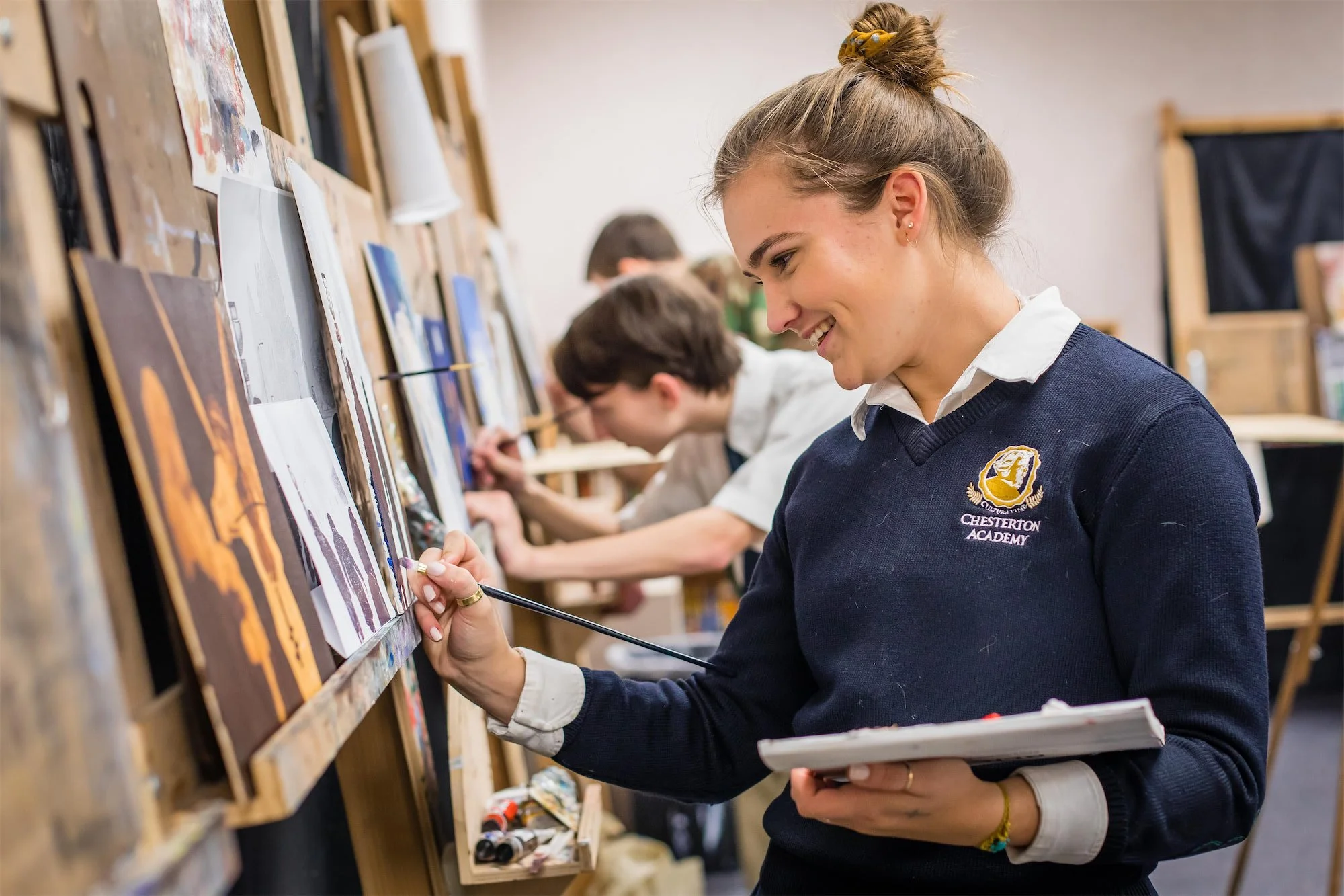 A young woman with a bun in her hair, wearing a navy sweater with a crest that says Chesterton Academy, is smiling while painting on a canvas in an art classroom with other students. She holds a sketchbook and a paintbrush, working at an easel.