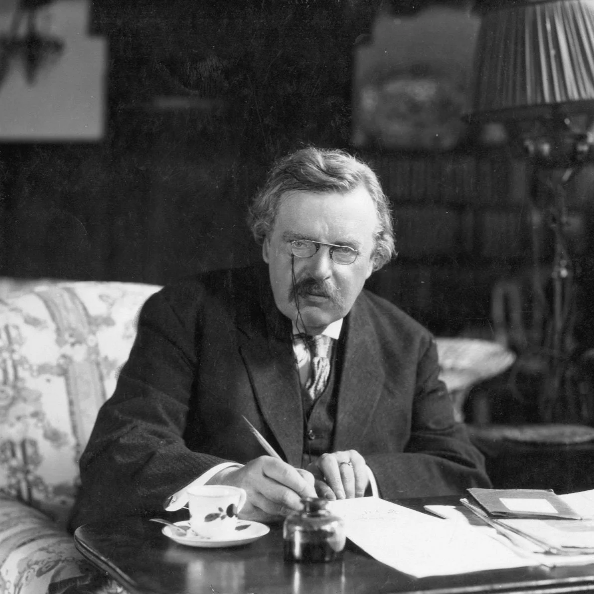 A black-and-white photo of a man with glasses, a mustache, and wavy hair, sitting at a table with papers, a teacup, and an ink bottle, in a room with bookshelves.