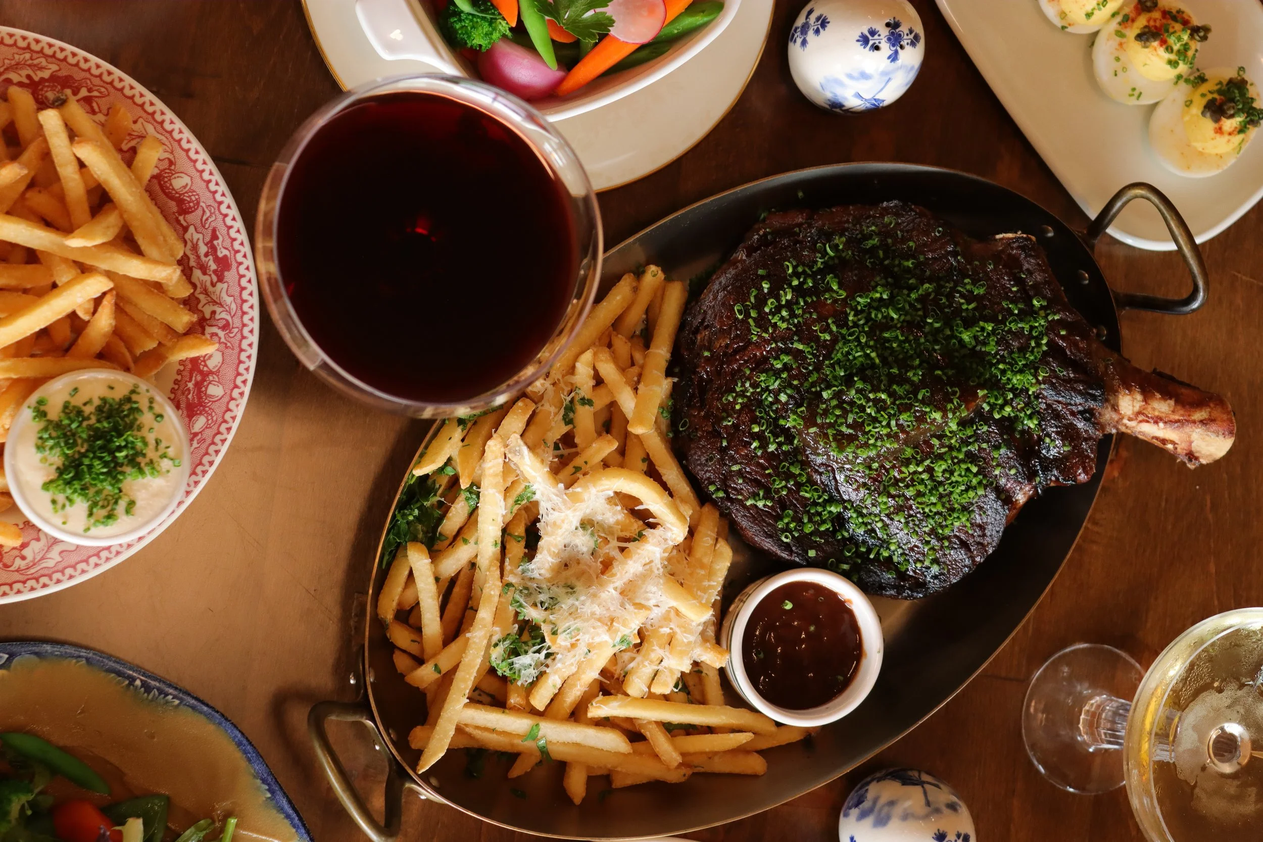 A table with various dishes including French fries, a glass of red wine, a cooked beef steak with chopped green onions, a salad with vegetables, deviled eggs with chili flakes, and small bowls of dipping sauces.