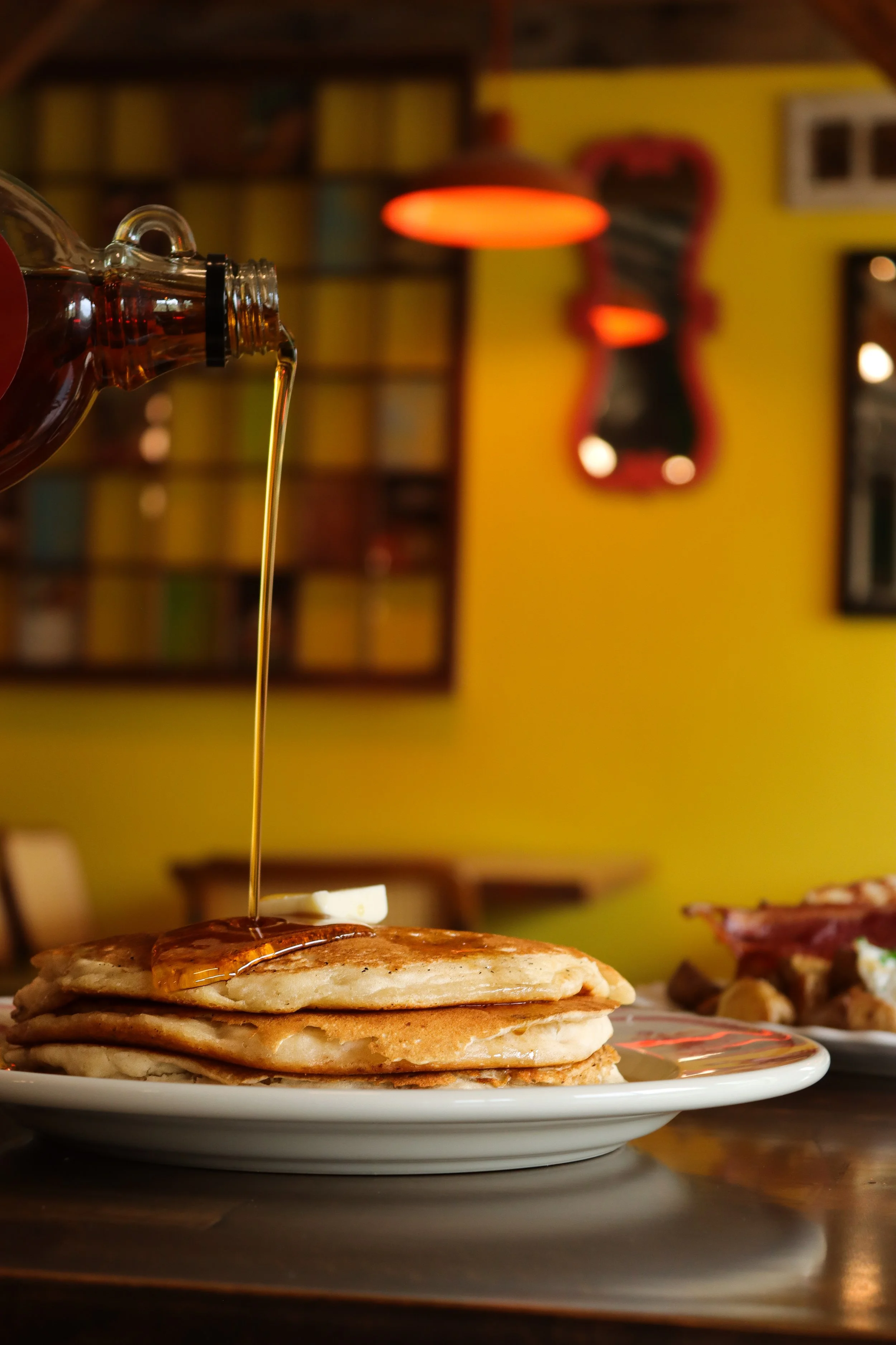 A stack of pancakes with syrup being poured on top, with a pat of butter melting on the pancakes, on a white plate in a cozy restaurant setting.