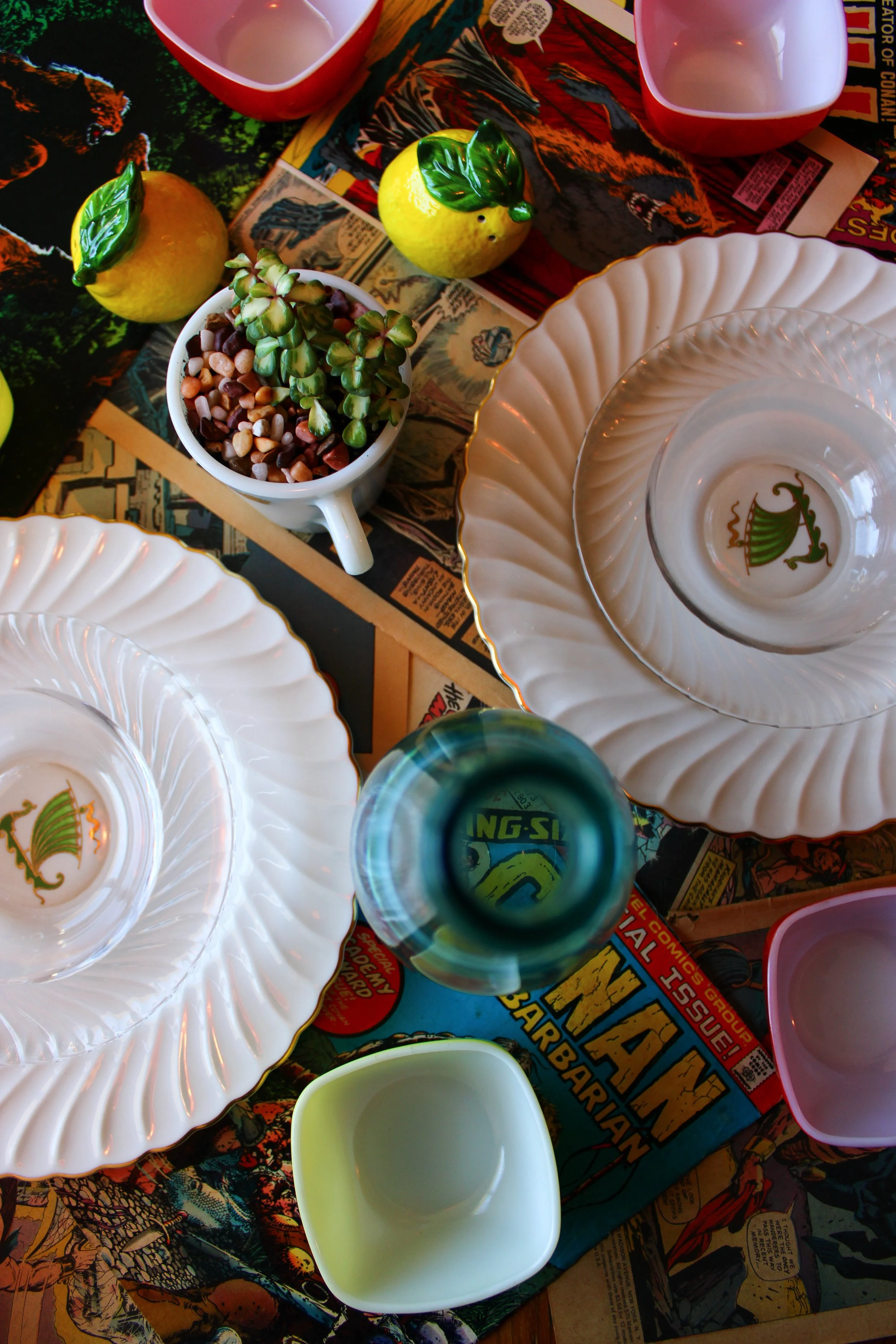 Colorful table setting with white ceramic plates featuring a green floral design, yellow and red lemon-shaped salt and pepper shakers, a small white pot with a plant, empty cups, and a comic book-themed tablecloth.