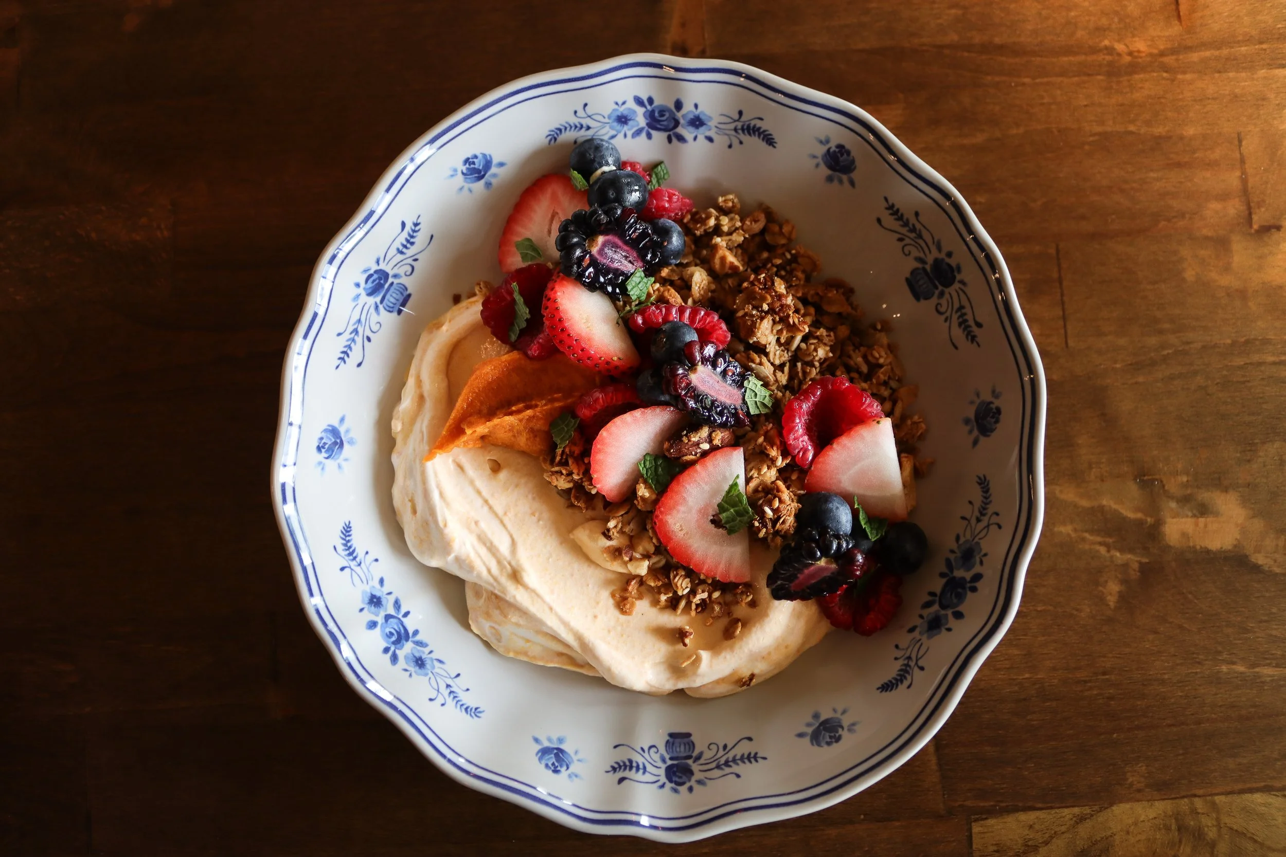 A decorative bowl with a blue floral pattern containing granola, sliced strawberries, blueberries, raspberries, and banana slices on a wooden surface.