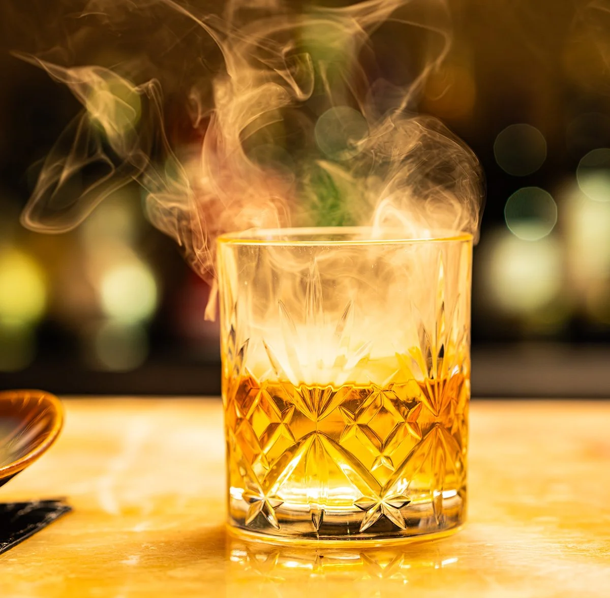 A glass of whiskey with smoke rising from it on a bar counter, with a blurred colorful background.