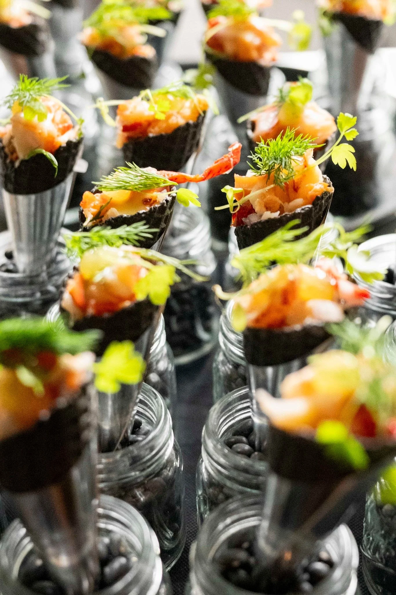 Miniature food appetizers in small glass jars, topped with seafood, herbs, and garnishes, arranged in a pattern.