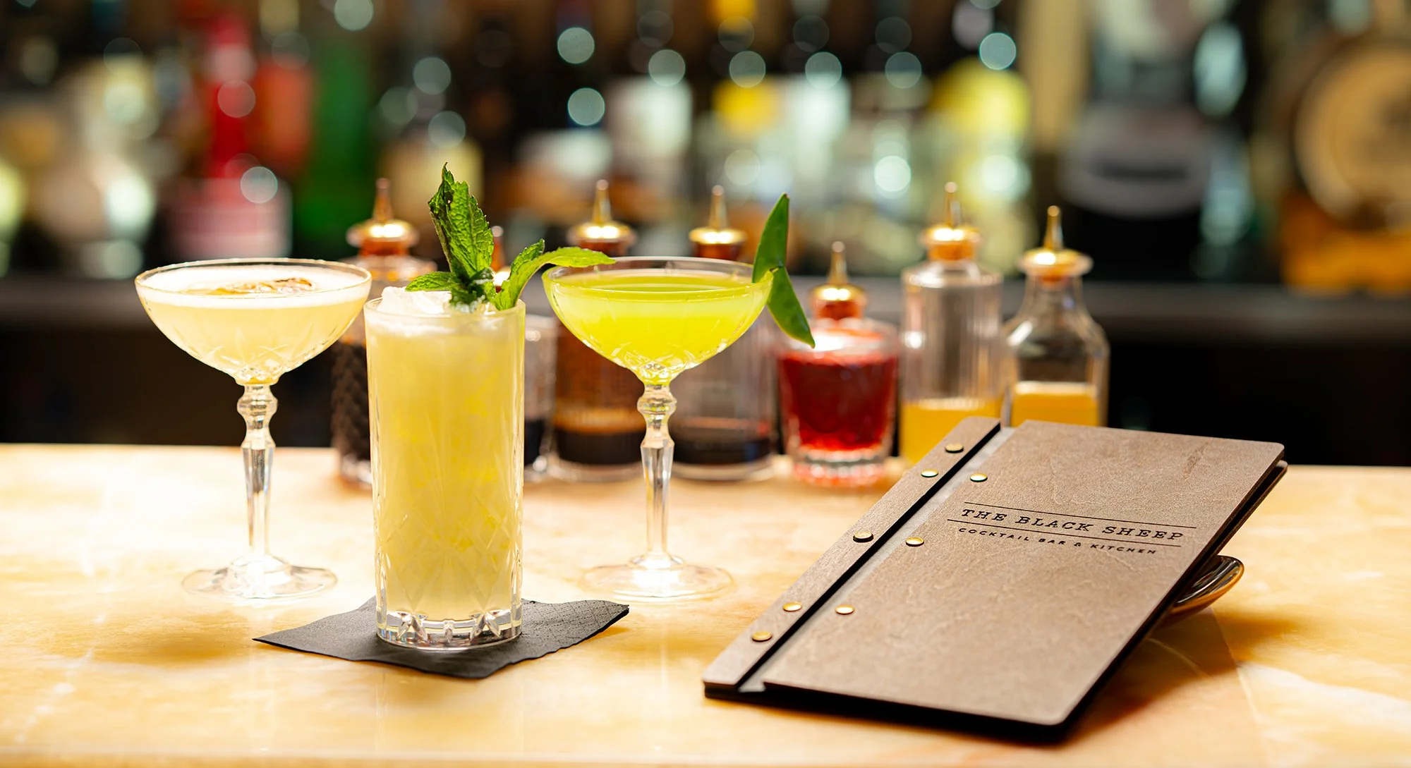 Three colorful cocktails with garnishes on a bar counter, with various bottles in the background, and a menu on the right side.