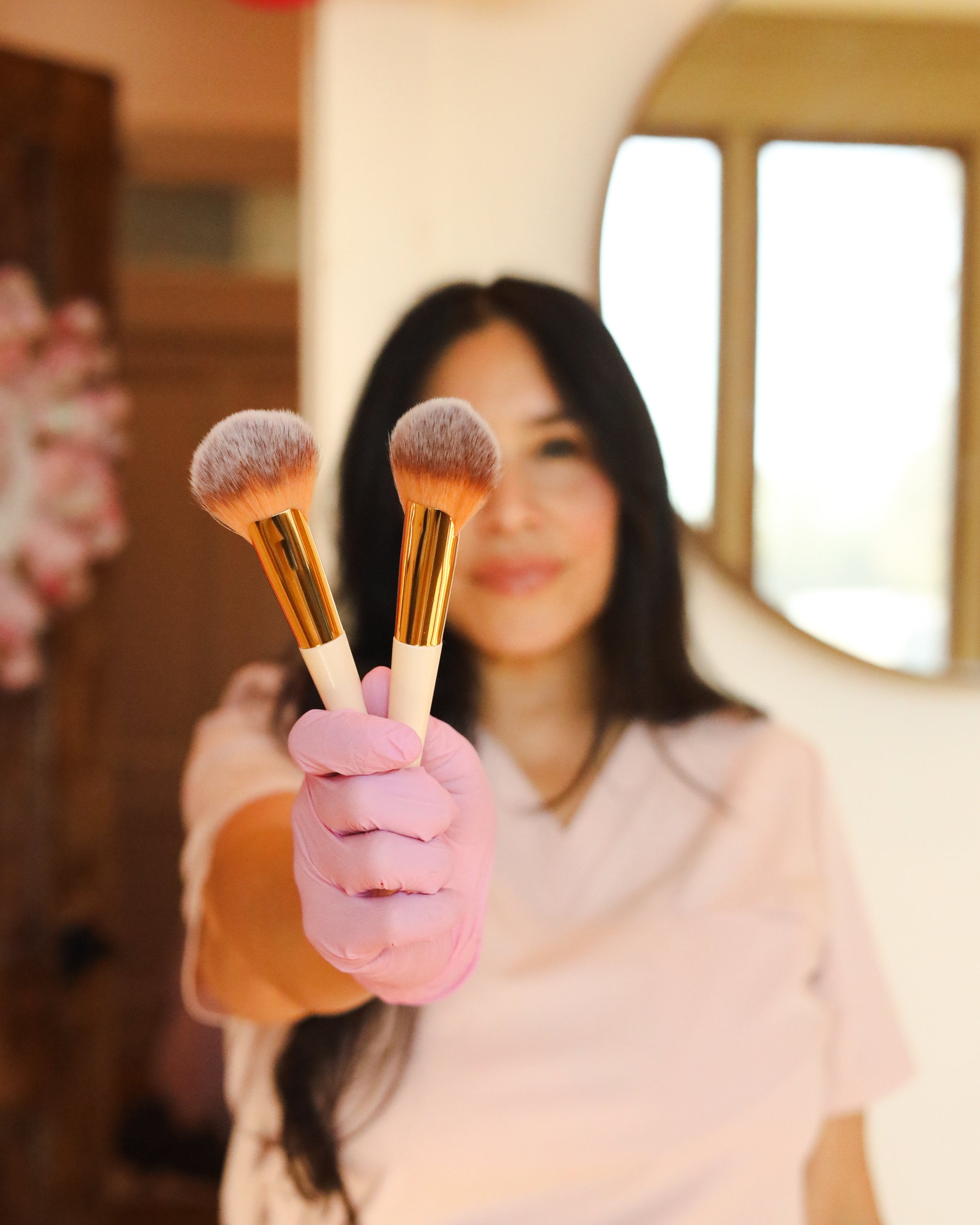 Woman smiling and holding two makeup brushes with pink gloves and gold handles, indoors near a window.