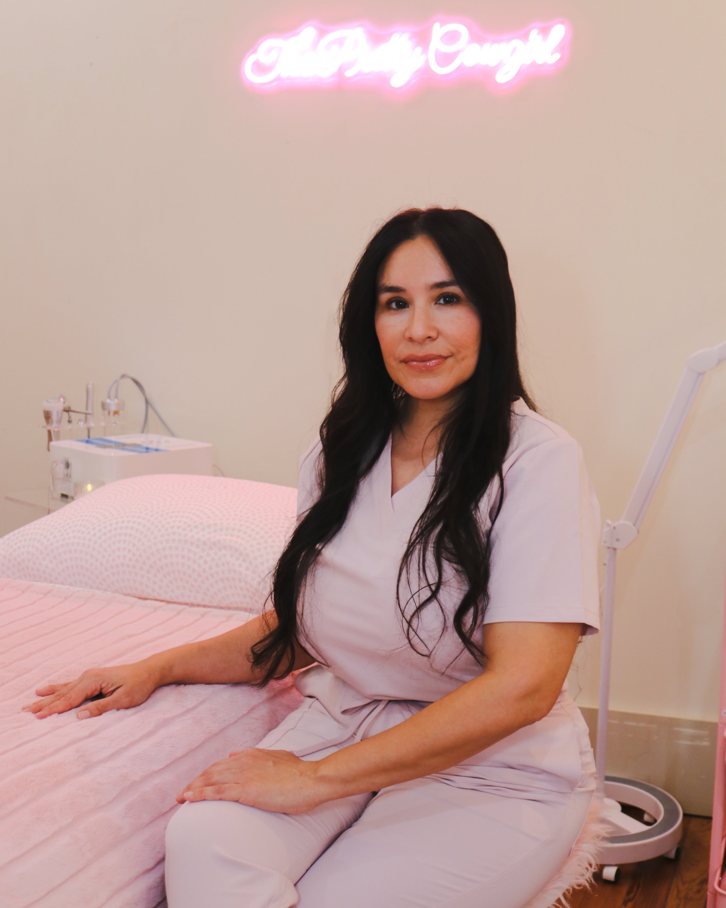 A woman in light pink scrubs sitting on a pink bed in a beauty or spa clinic, with a pink neon sign above her that reads 'The Pinky Couch'. There is medical or beauty equipment on a table behind her.