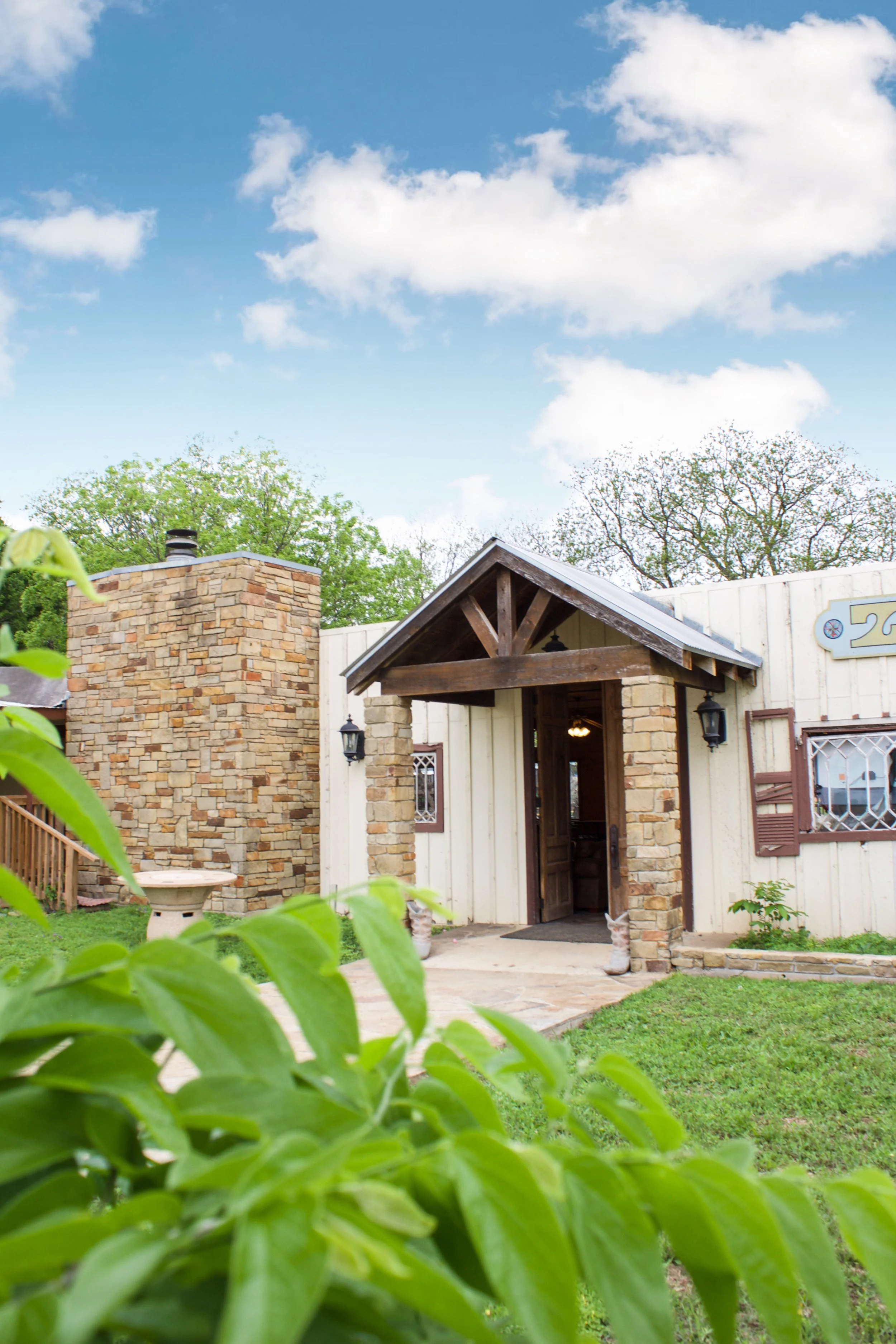 Front view of a house with a stone chimney, a brick and wood entrance, lush green lawn, and leafy plants in the foreground under a blue sky with scattered clouds.