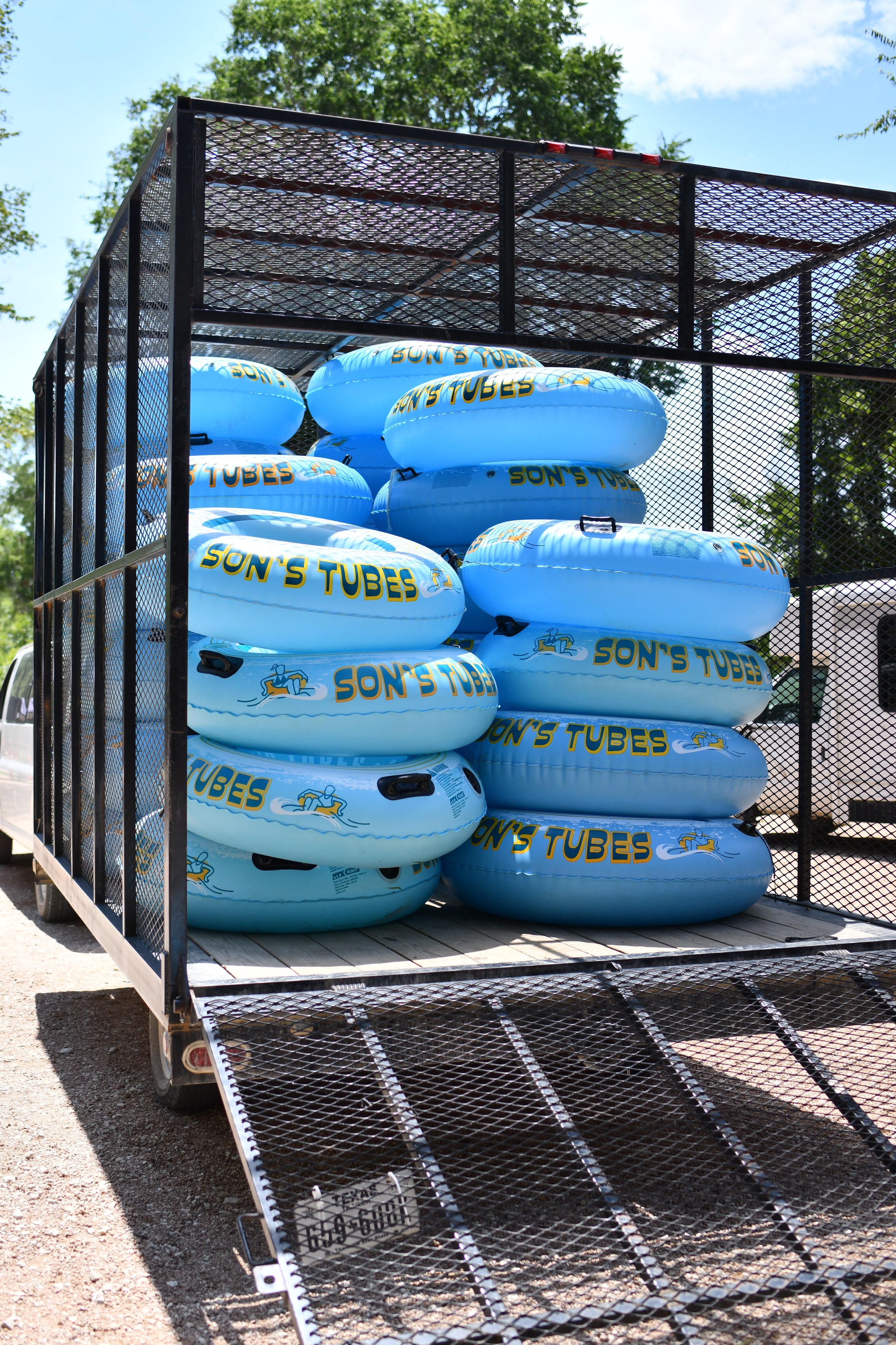 Stacked blue inner tubes labeled 'Son's Tubes' in a black metal trailer with a ramp, outdoors on a sunny day with trees in the background.