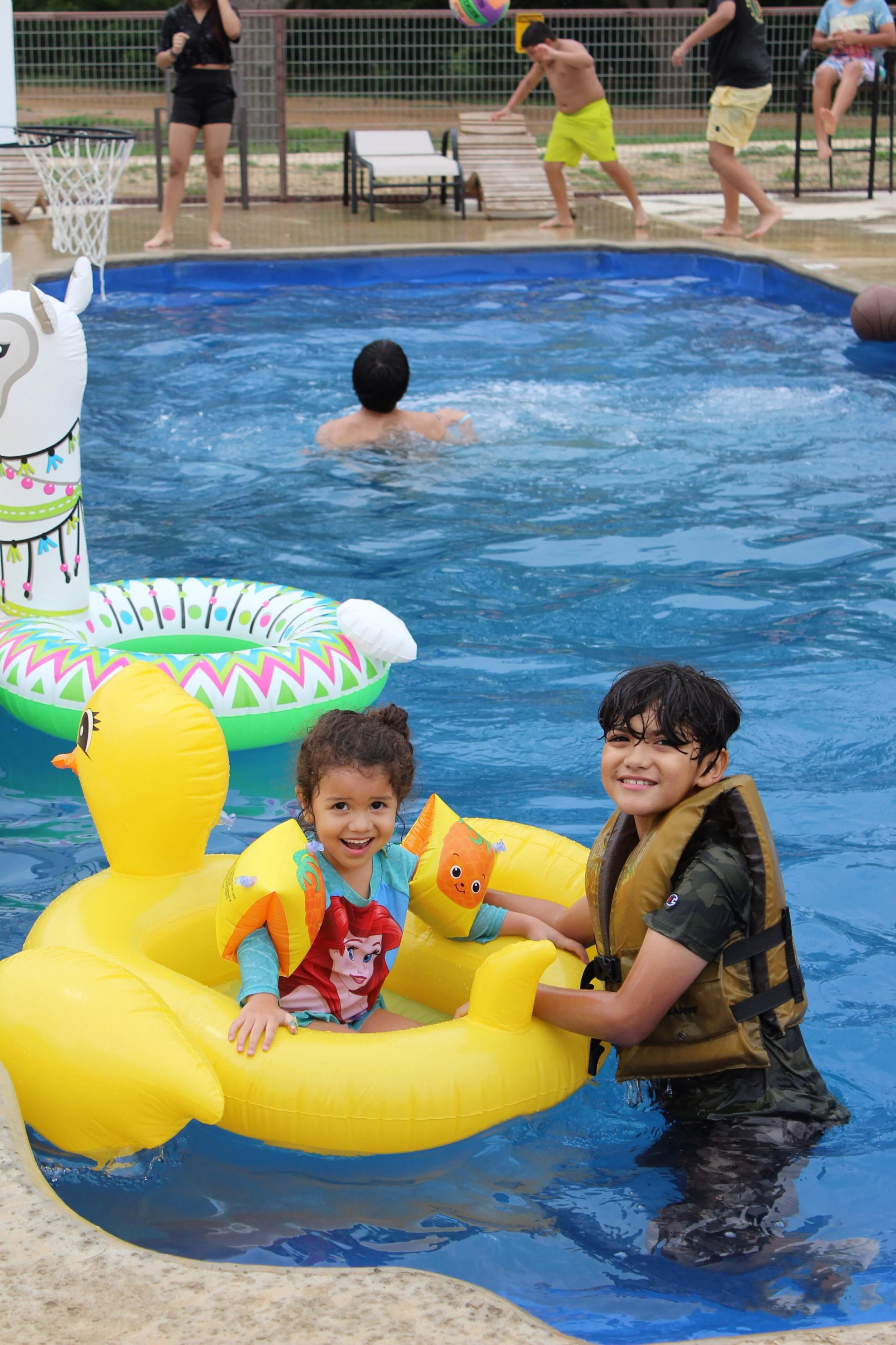 A boy and girl in a yellow inflatable duck float smiling in a swimming pool, with other children swimming and playing in the background.