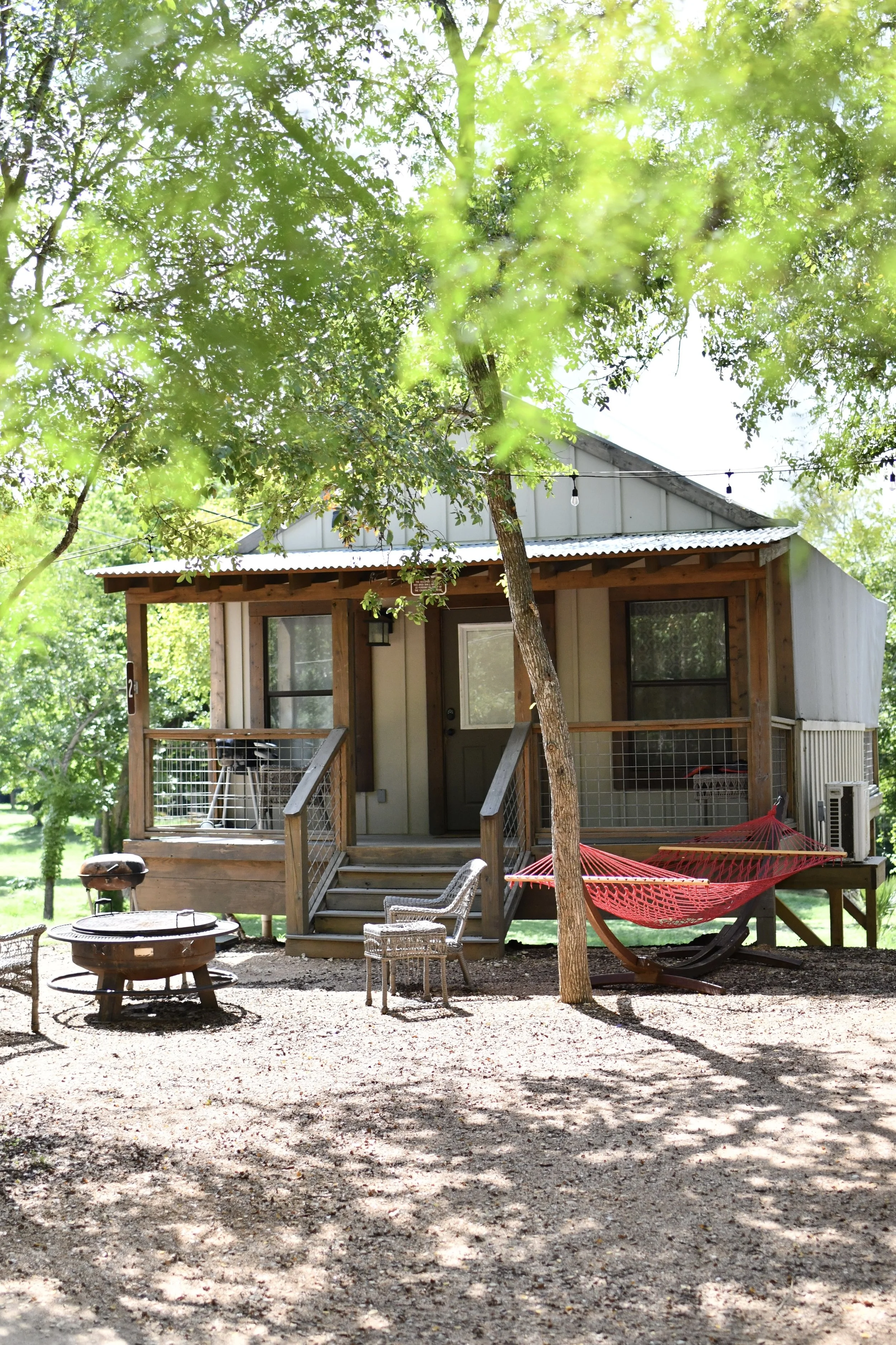 A small wooden house with a porch, surrounded by trees, with a hammock, chair, fire pit, and grill outside.