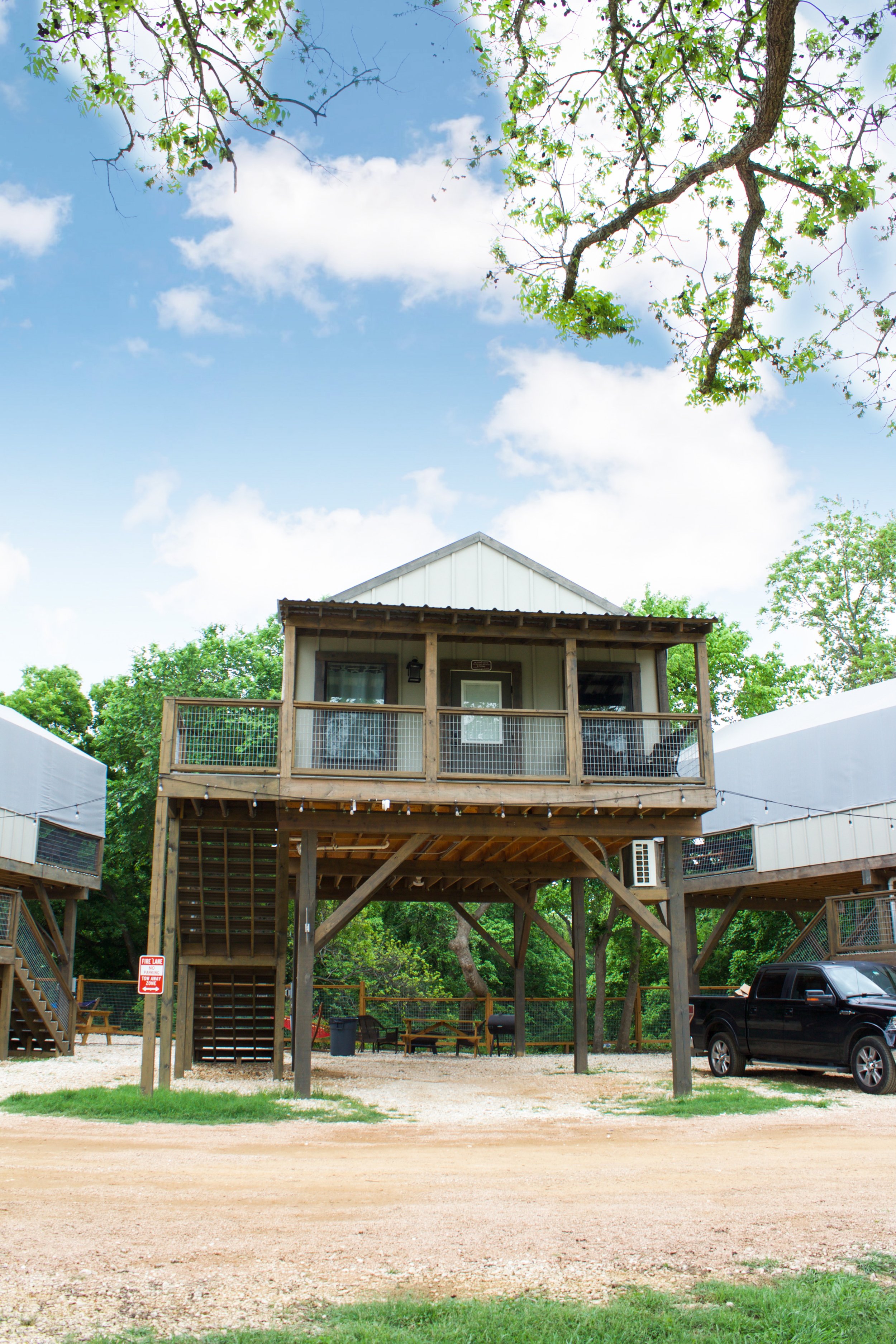 A raised house with wooden stairs and a deck, surrounded by trees, with a black truck parked underneath, and a clear blue sky with clouds overhead.