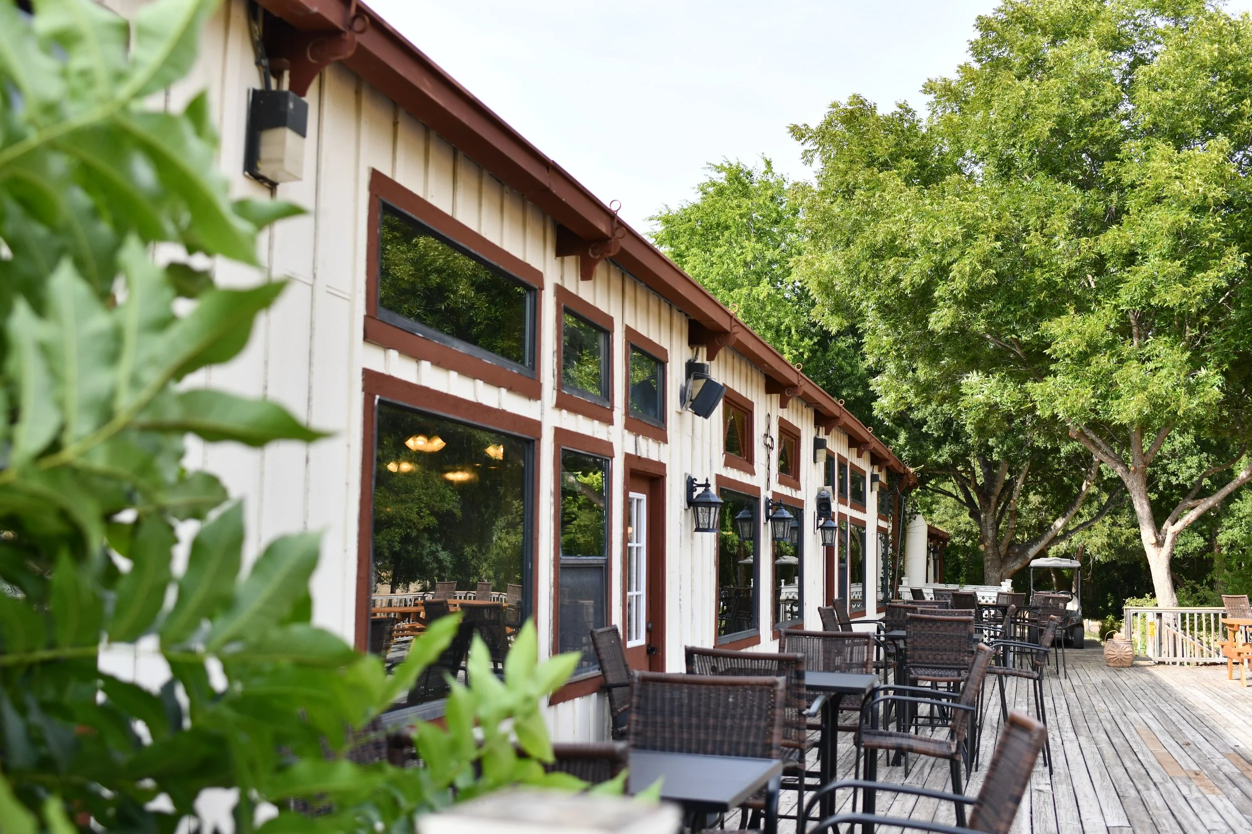 Outdoor patio of a restaurant or cafe with multiple tables and chairs, a building with several windows and exterior-mounted lights, surrounded by lush green trees.