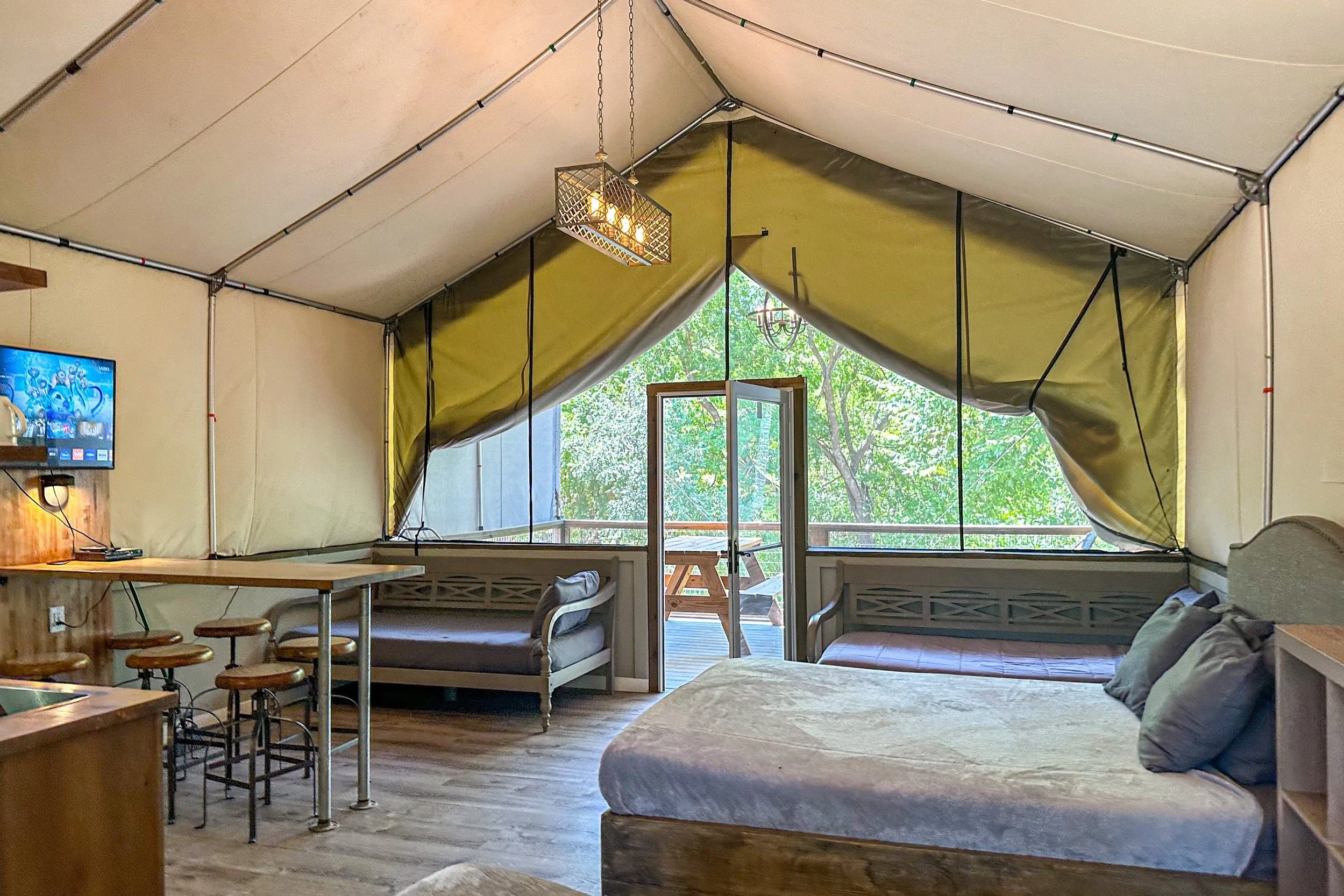 A cozy tent interior with a bed, a small sofa, a dining area with stools, a wall-mounted TV, and large open doors leading to a wooden deck with outdoor furniture, surrounded by trees.