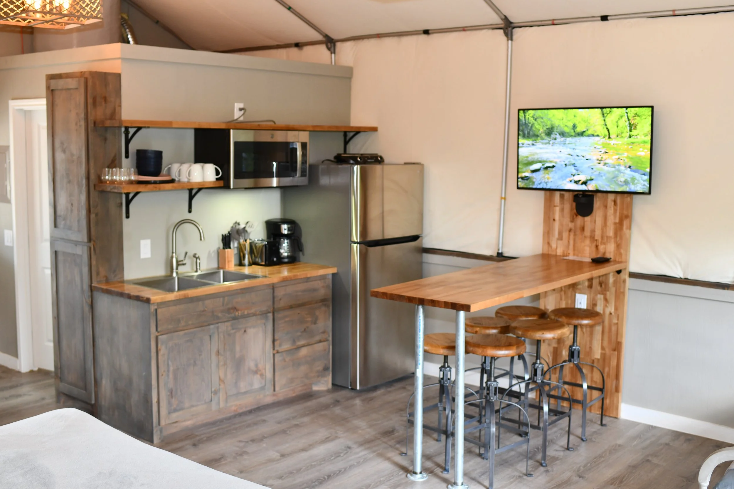 Small kitchen area with wooden cabinets, countertop, microwave, refrigerator, coffee maker, and open shelves with cups and glasses. Adjacent to a wooden bar table with four stools, and a wall-mounted flat-screen TV showing a nature scene.