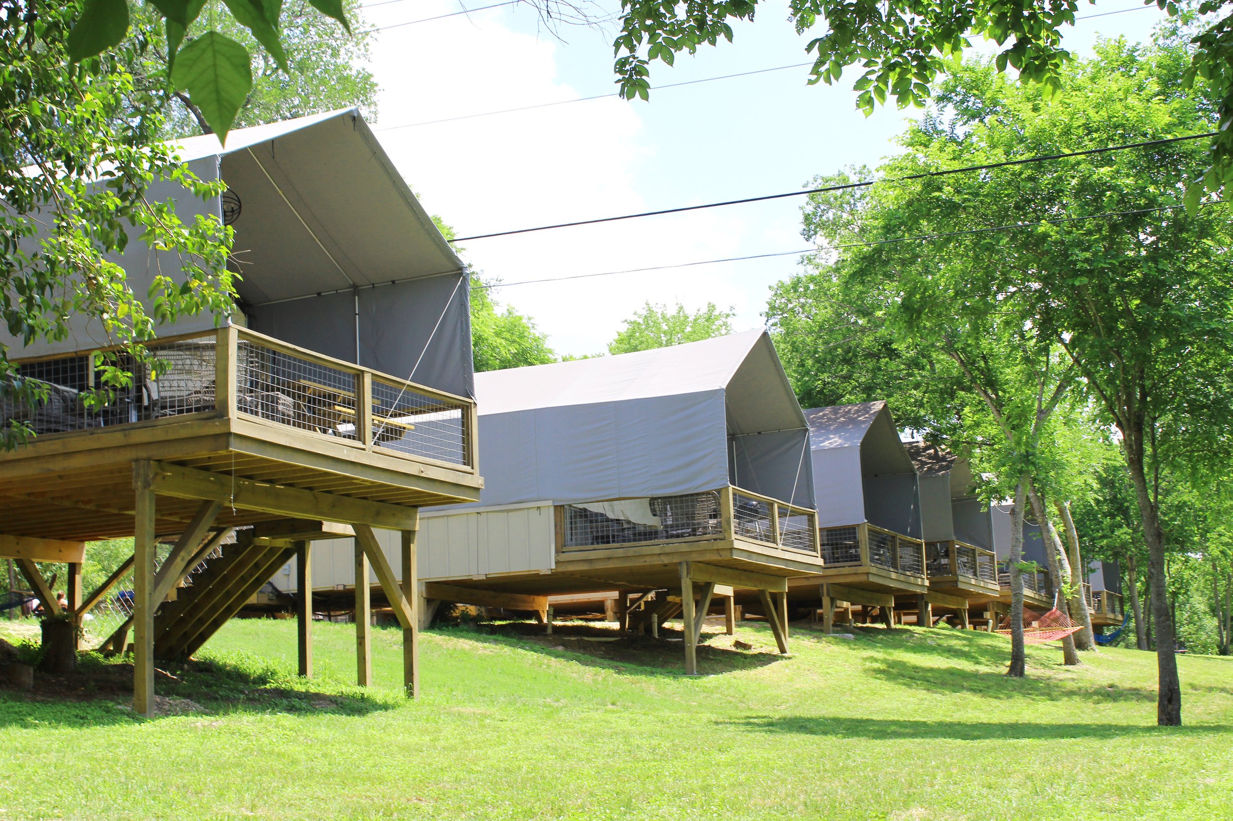 Row of elevated tents on wooden platforms in a green park with trees, blue sky, and sunlight