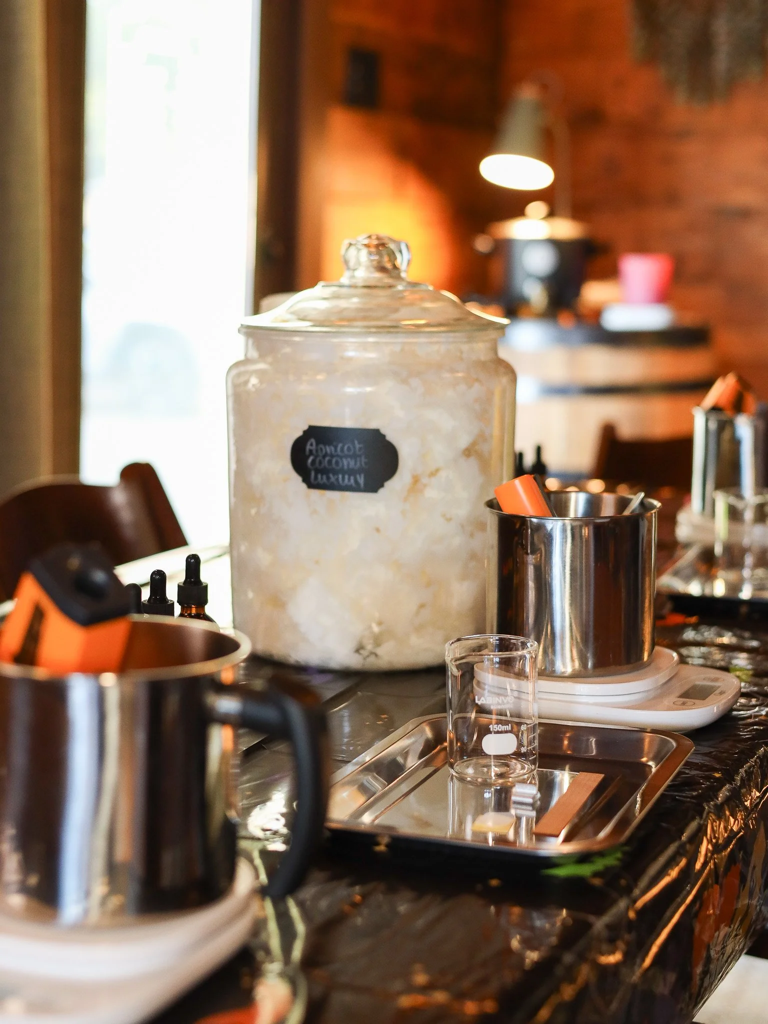 A large jar labeled 'Amiot Coconut Luxury' surrounded by equipment like a graduated cylinder, small bottles, and electronic scales on a black tablecloth in a warmly lit, rustic indoor setting.
