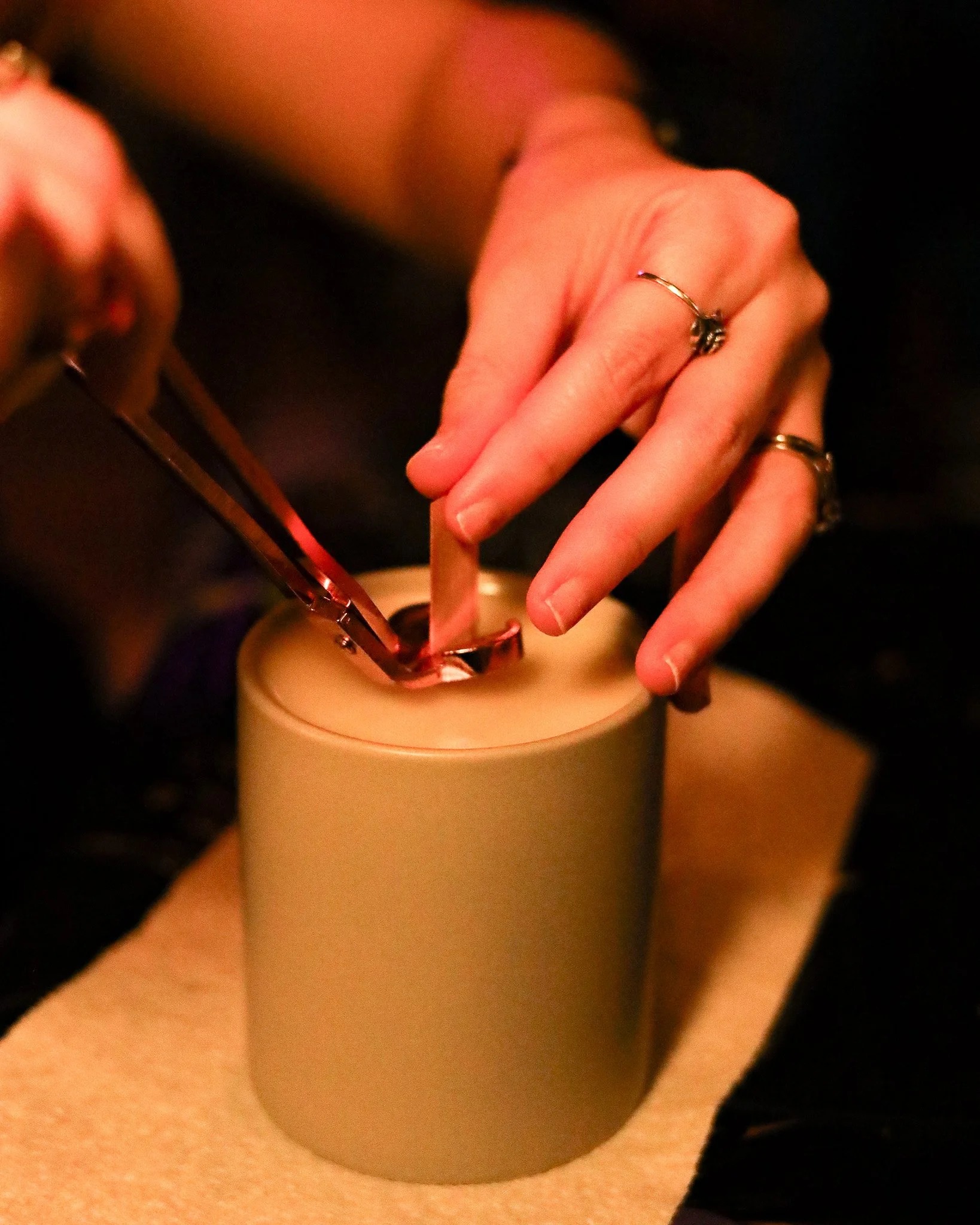 A person's hands are seen placing a piece of soap into a cylindrical white container, with one hand holding a pair of tongs and the other stabilizing the container. The person is wearing multiple rings on fingers.