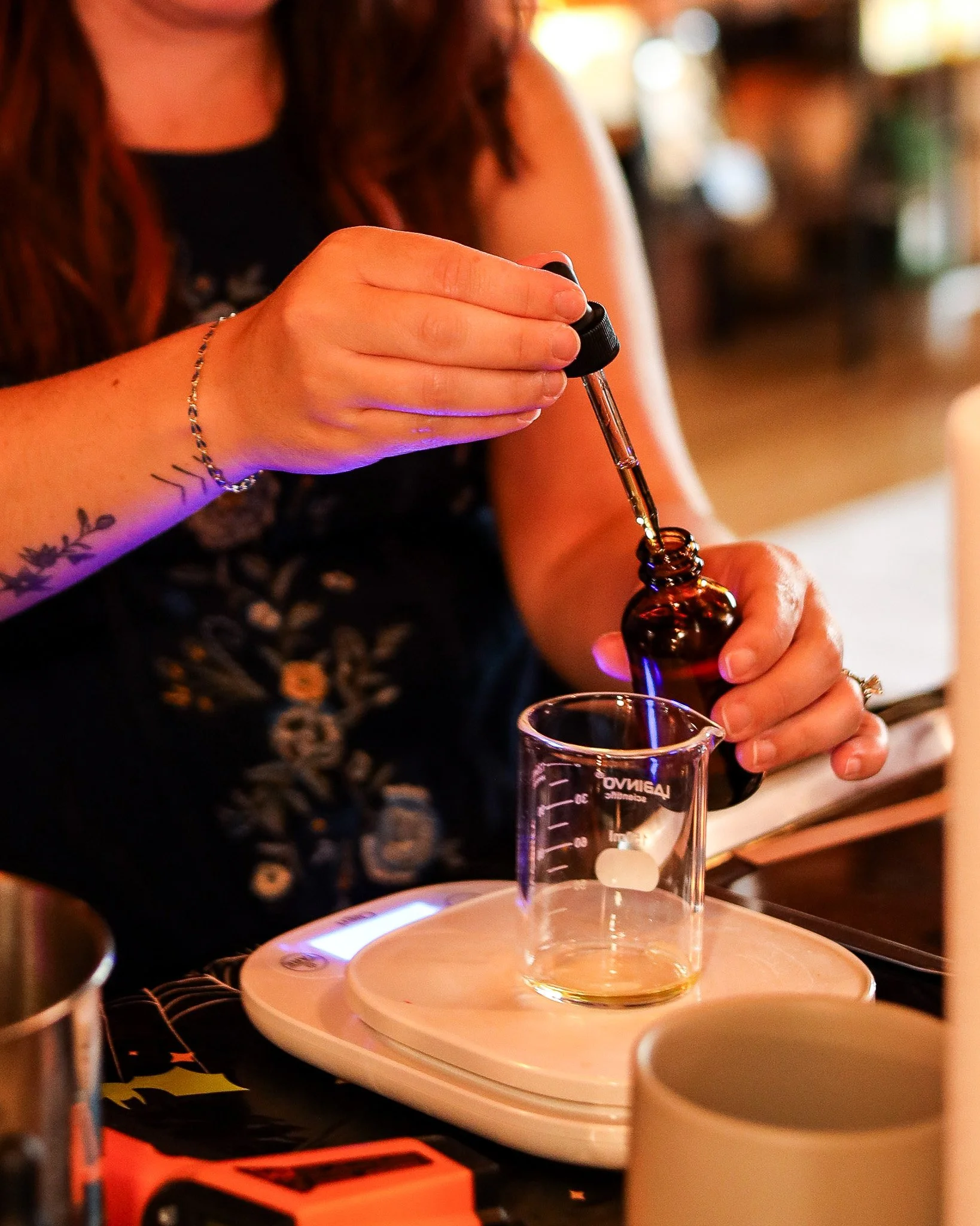 A woman using a dropper to transfer liquid into a glass beaker on a digital scale in a laboratory or workshop.
