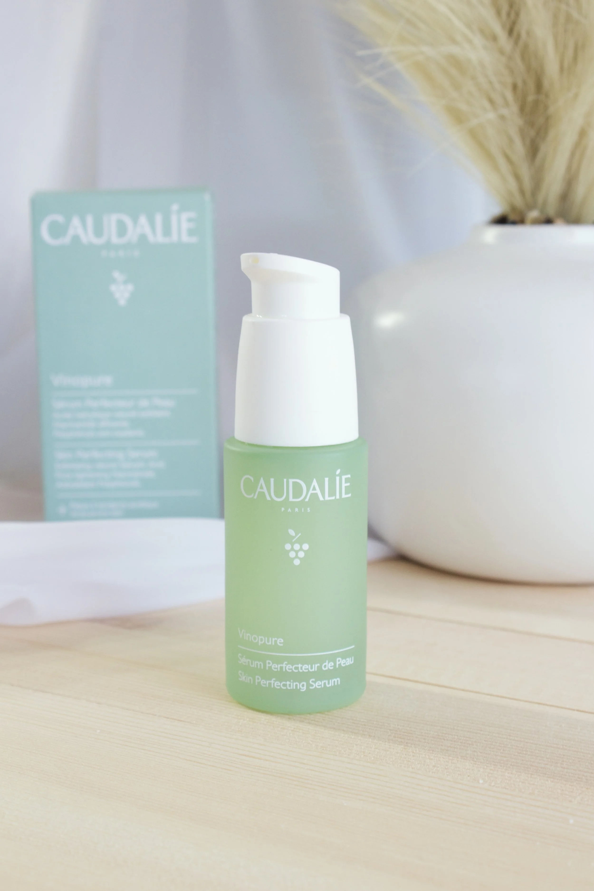 A green bottle of Caudalie Vinopure Skin Perfecting Serum on a wooden surface with a white vase and dried plants in the background.