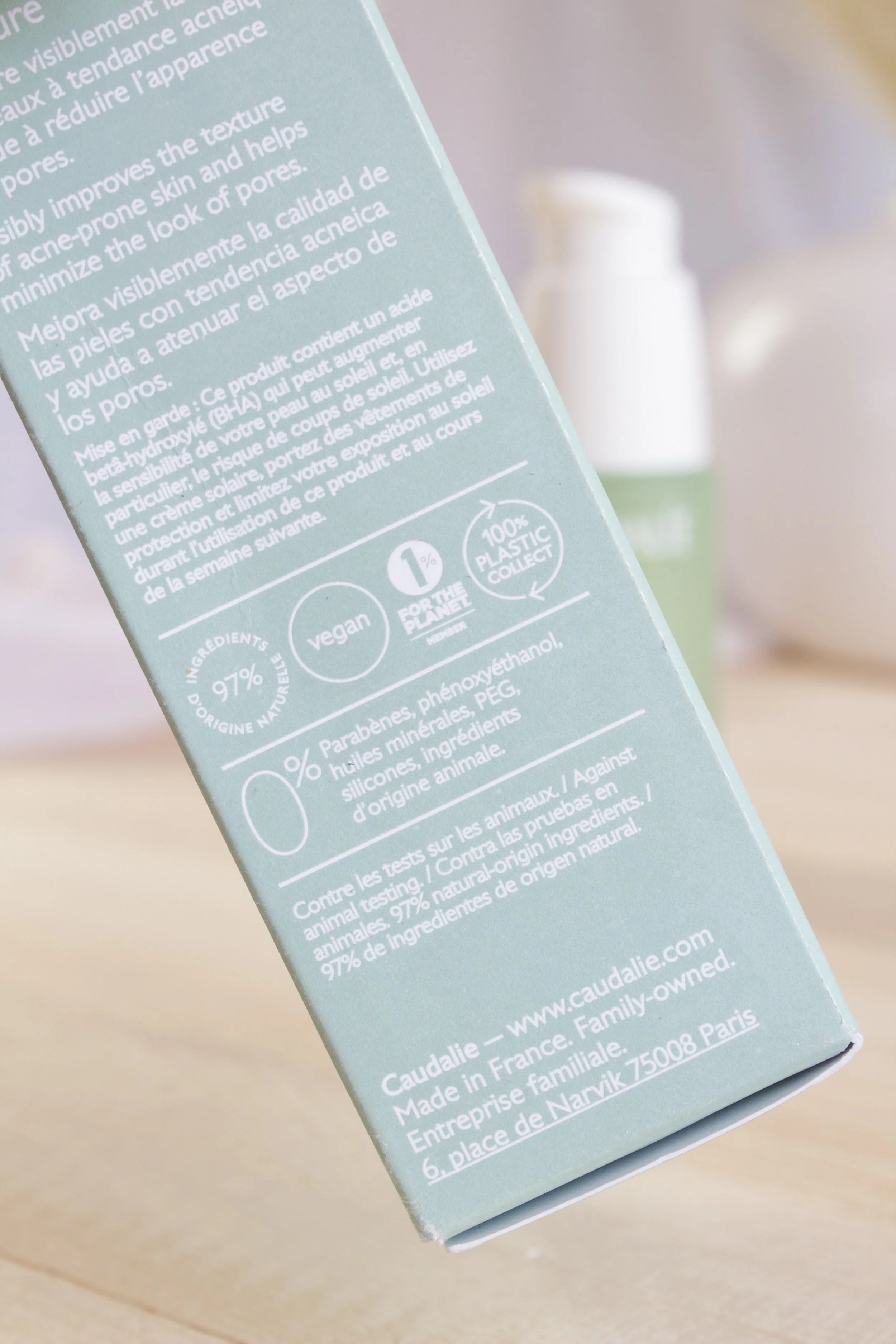 Close-up of a light blue skincare product box with text in French and English, alongside a white and green bottle in the background.