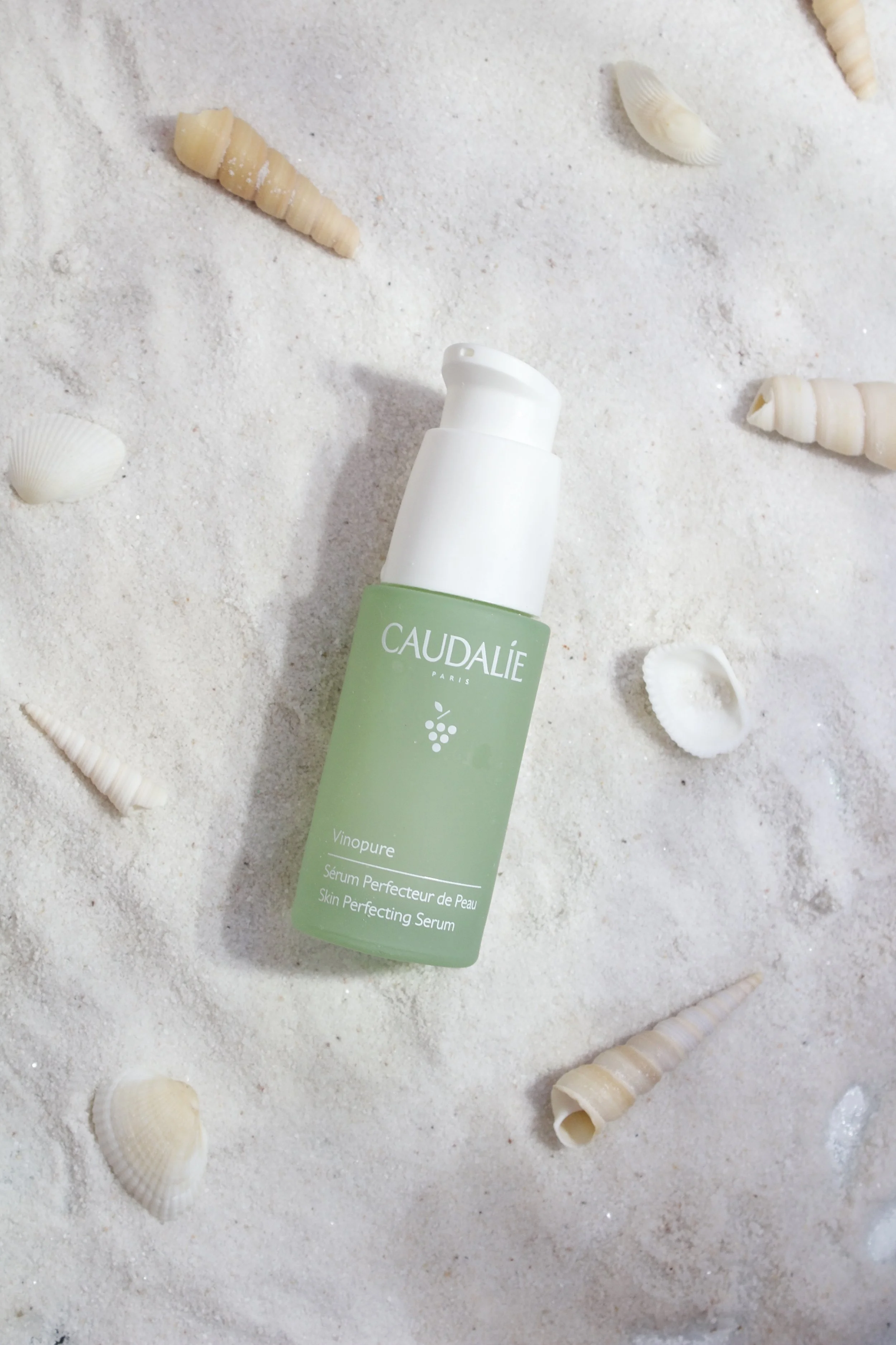 A green bottle of Caudalie Vinopure Skin Perfecting Serum lying on white sand, surrounded by seashells.