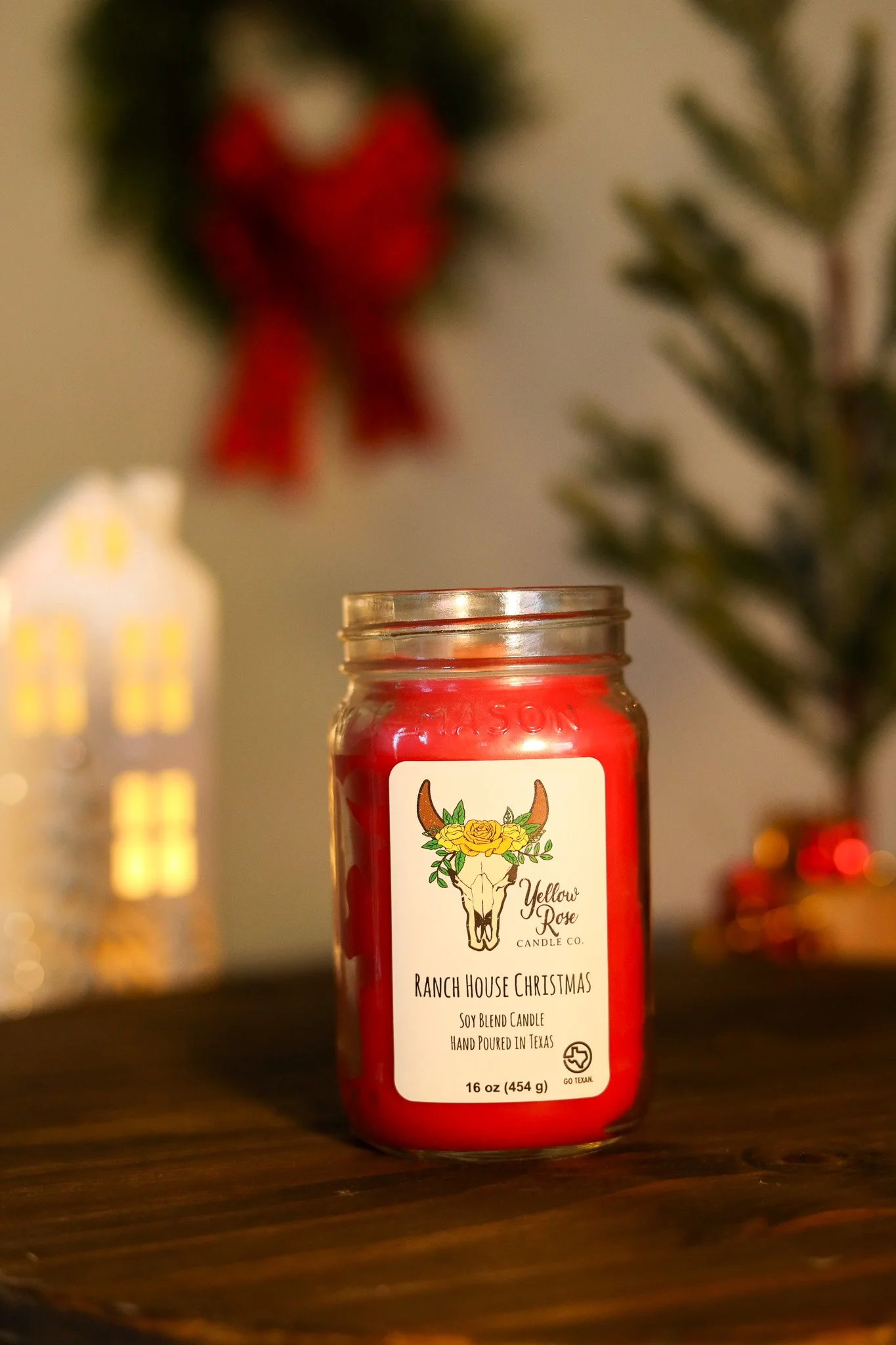 Red candle in a glass jar labeled Yellow Rose Ranch House Christmas, soy blend candle, hand poured in Texas, 16 oz, with blurred holiday decorations in the background.
