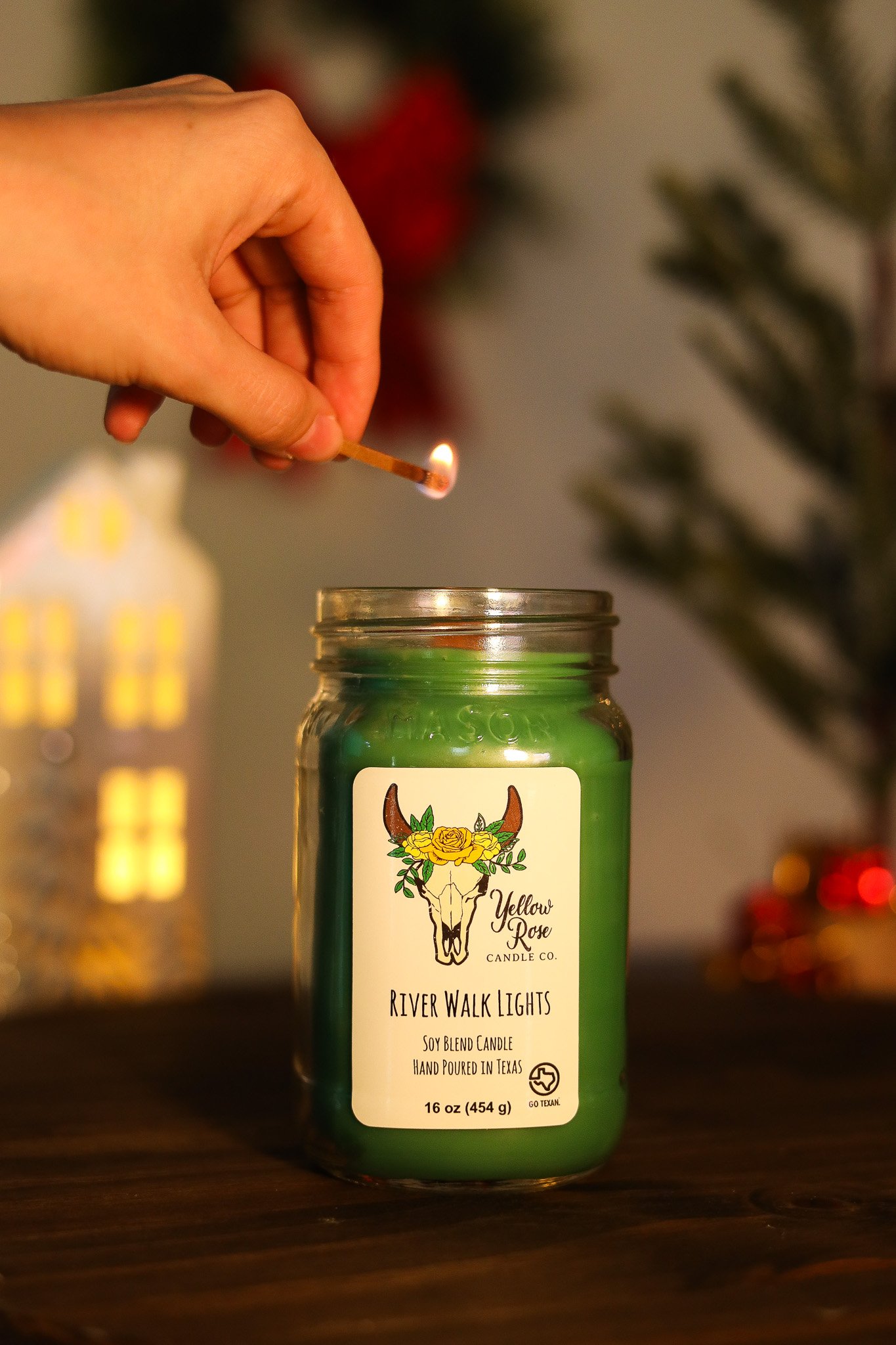 A hand holding a lit match above a green candle in a jar, with Christmas decorations in the background.