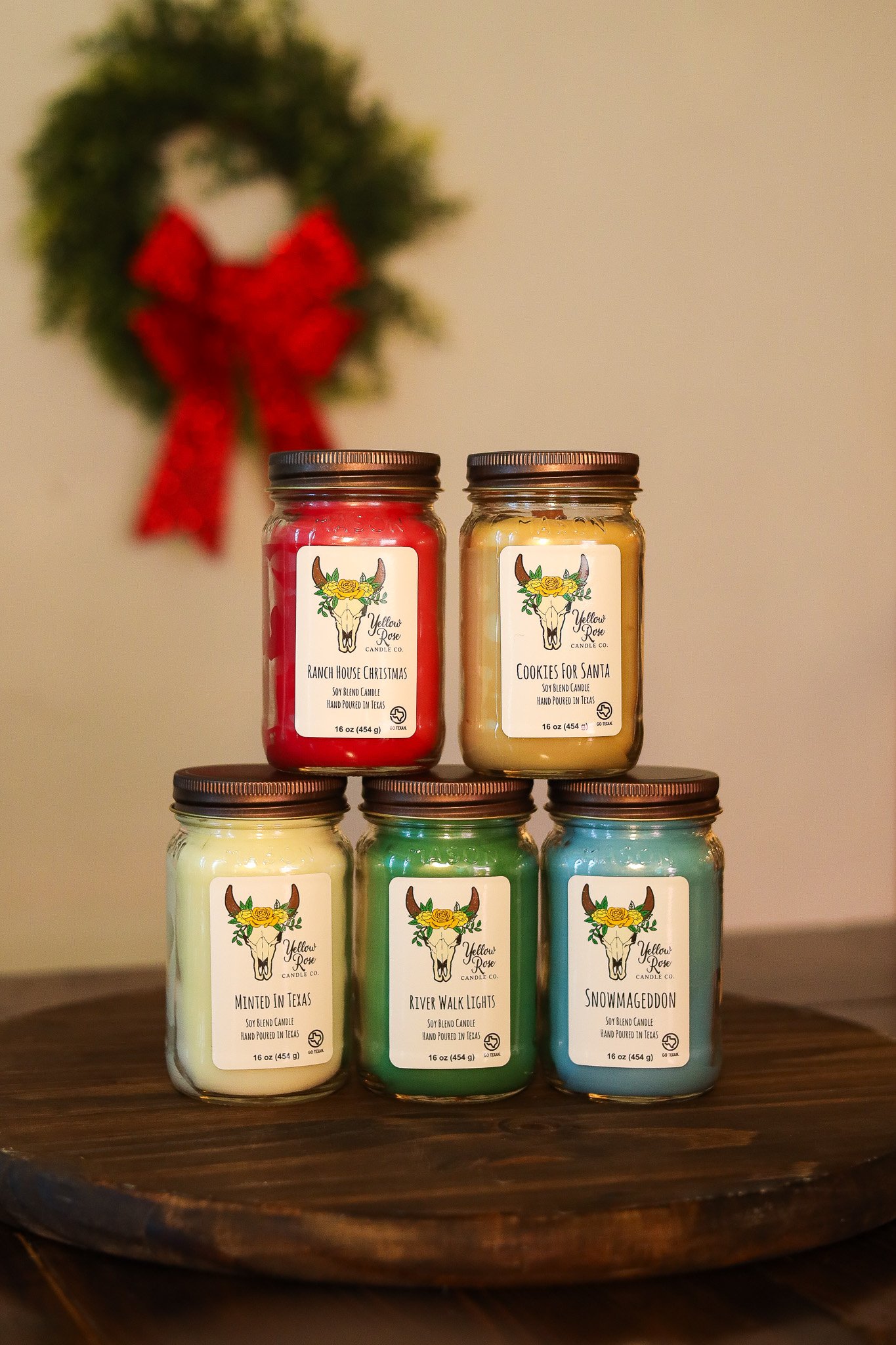 Six scented candles in glass jars with brown lids arranged in a pyramid on a wooden surface, with a holiday wreath and red bow in the background.