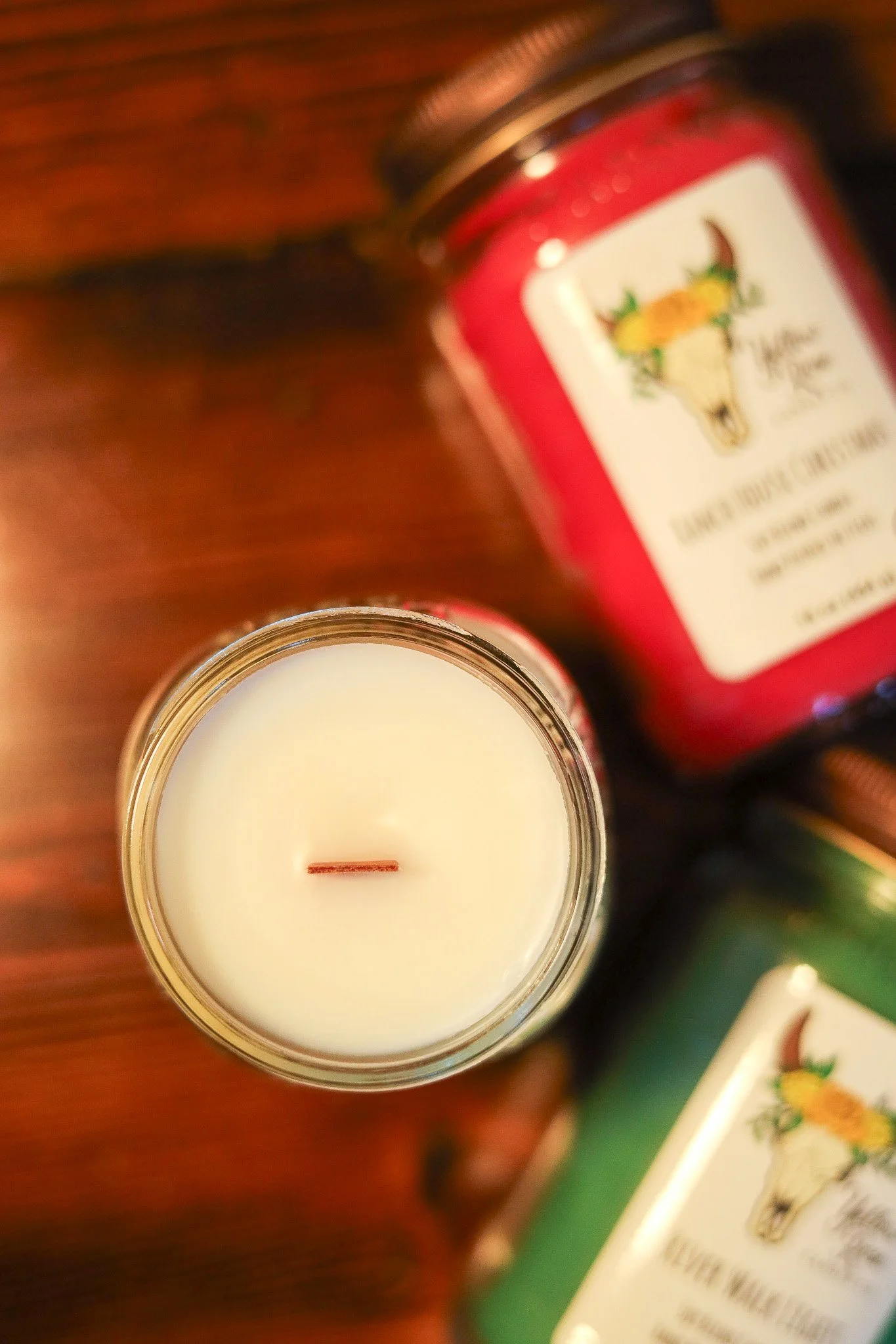 Close-up of a white candle in a glass jar, viewed from above, with a beard-shaped wick. In the background, there are candles in red and green jars with illustrations of a skull with horns and flowers.